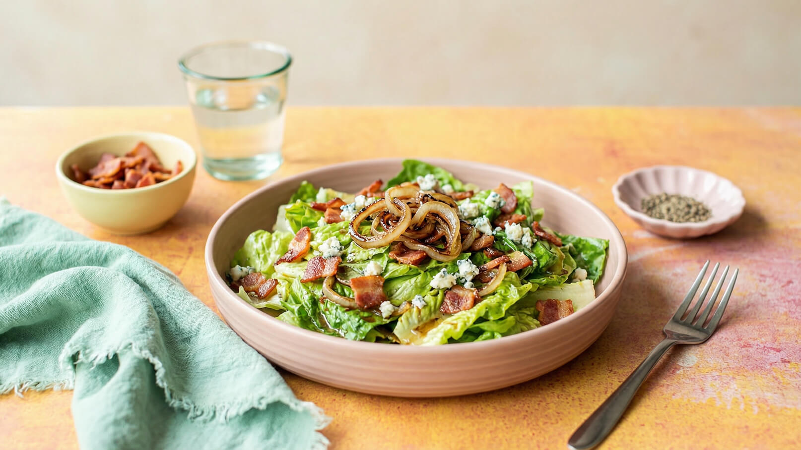 Eine Schüssel mit knackigem Salat, garniert mit knusprigem Speck, gebratenen Zwiebeln und zerbröckeltem Käse. Im Hintergrund befinden sich ein Glas Wasser und eine kleine Schüssel mit Speckstücken. Der Tisch hat eine warme, orangefarbene Oberfläche.