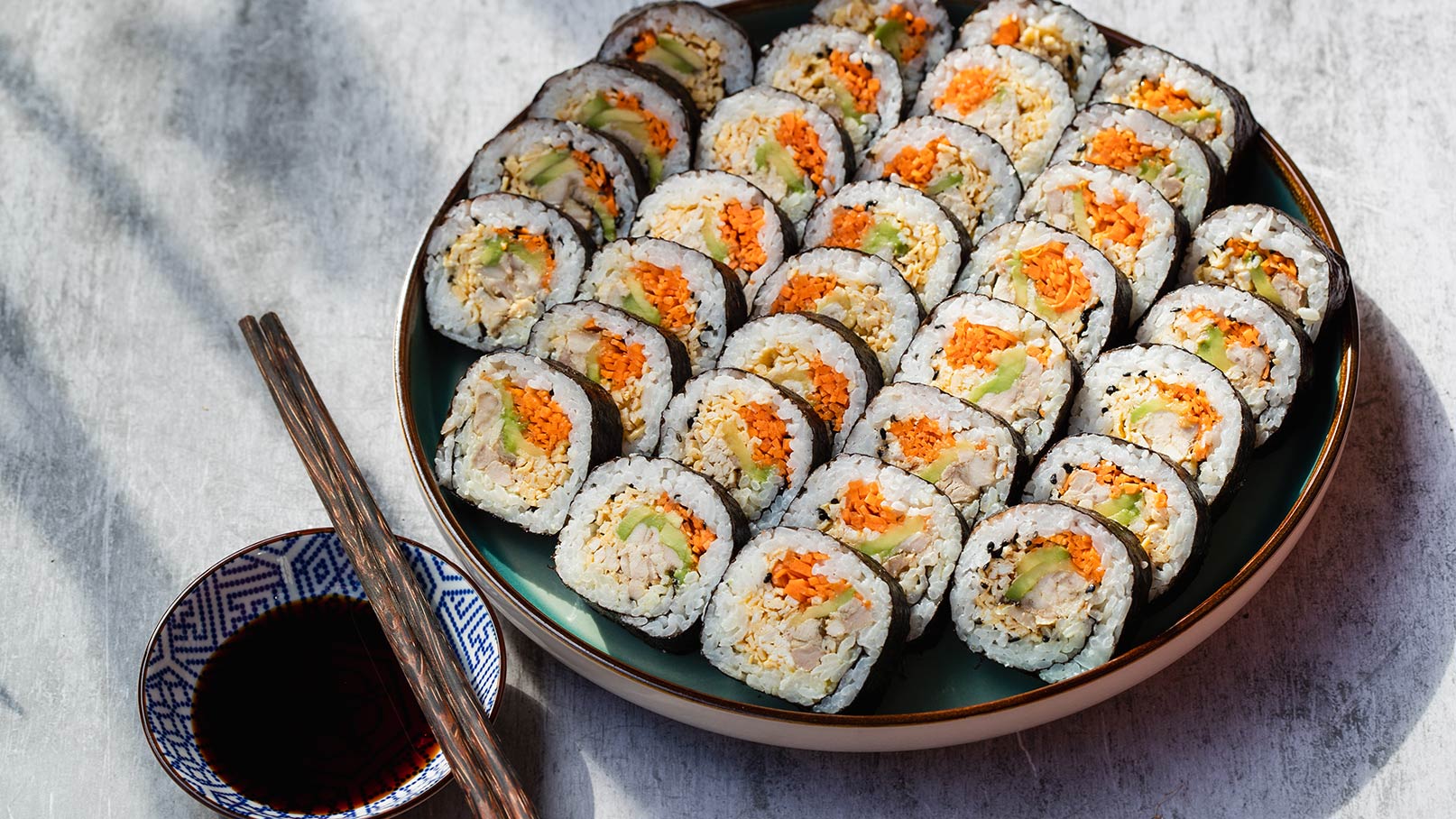 Ein Teller mit mehreren Sushi-Rollen, die mit Reis, Seealgen und bunten Füllungen wie Karotten und Gemüse gefüllt sind. Neben dem Teller liegen Essstäbchen und eine kleine Schüssel mit Sojasauce.