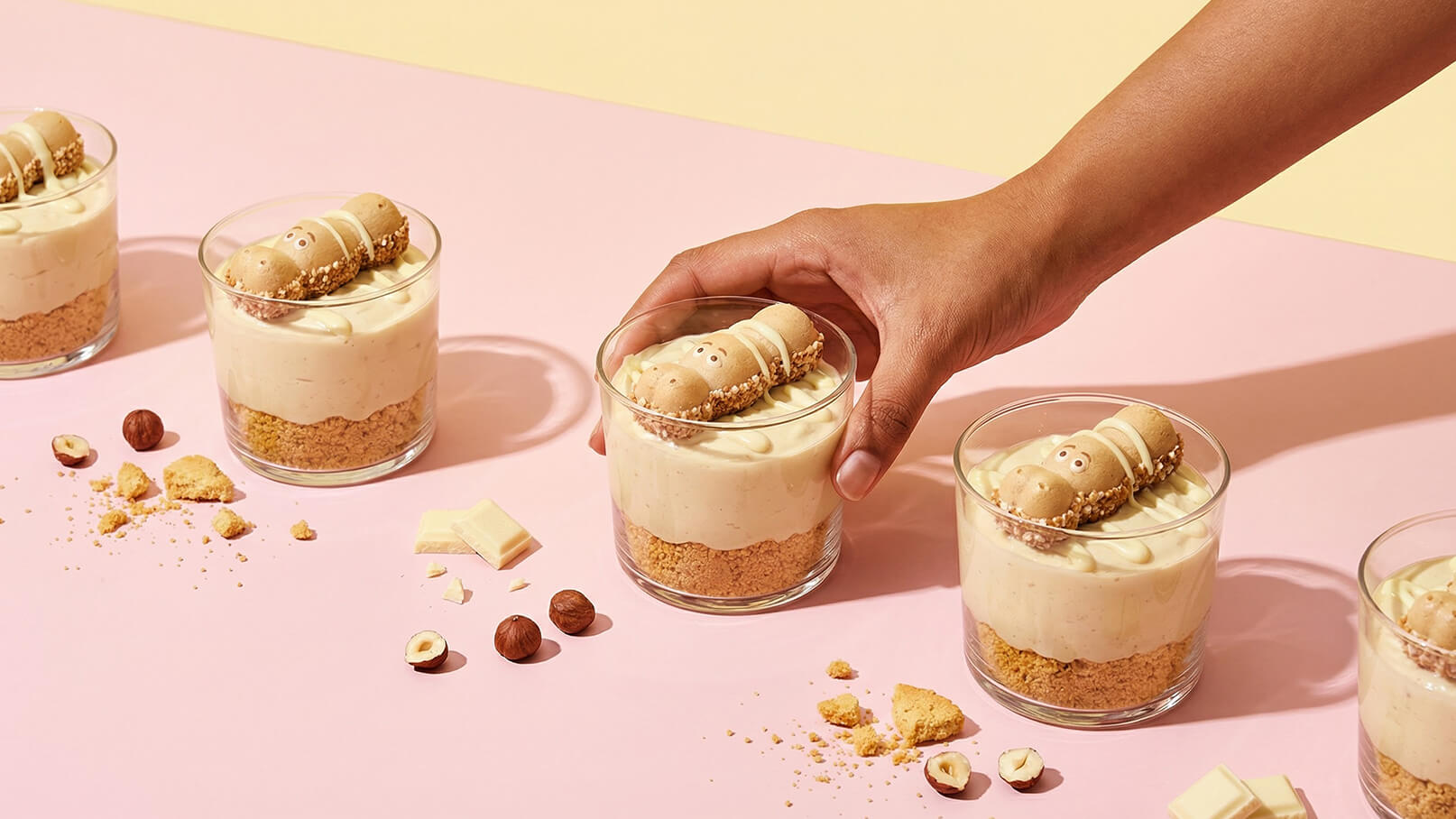 Eine Hand greift nach einem Glas mit einem Dessert, das aus einer cremigen Schicht und einem Boden aus Keks besteht. Auf dem Dessert liegt eine runde Kekssorte. Im Hintergrund sind mehrere ähnliche Gläser mit Desserts angeordnet.