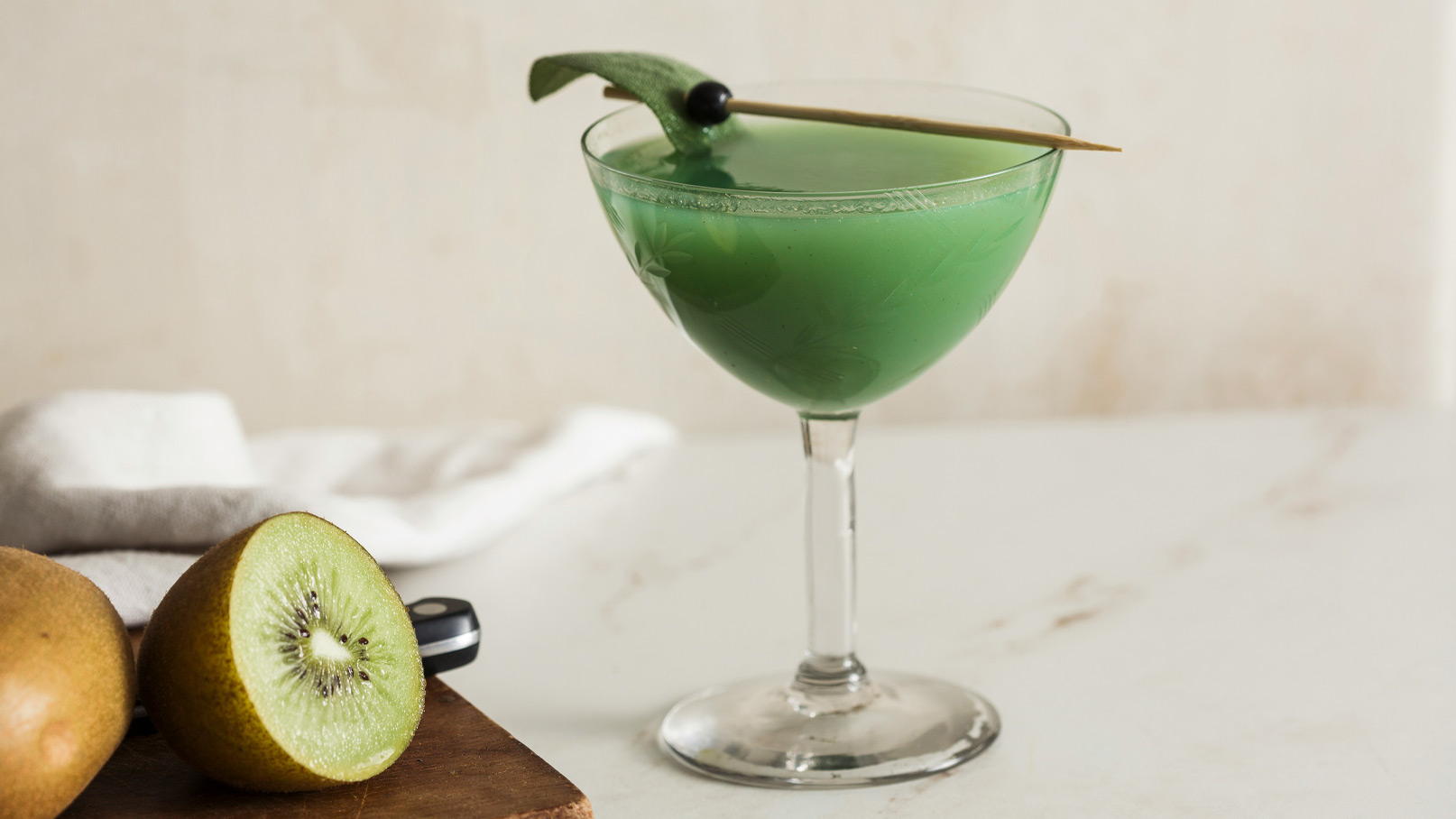 Ein grüner Cocktail in einem eleganten Glas, dekoriert mit einem Blatt und einer Olive auf einem Holzbrett. Neben dem Glas liegt eine halbierte Kiwi, die den frischen und fruchtigen Charakter des Getränks unterstreicht.