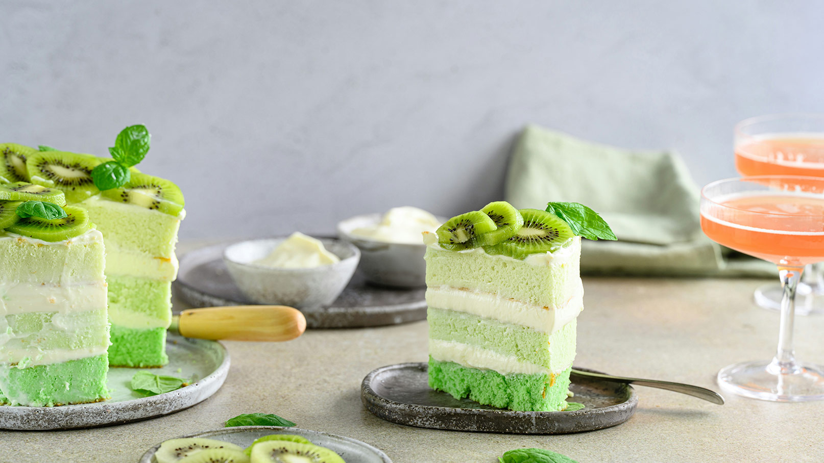 Ein Stück grüner Kuchen mit mehreren Schichten, dekoriert mit Kiwis und Minzblättern. Neben dem Kuchen befinden sich kleine Schalen mit Sahne und ein Löffel. Im Hintergrund stehen zwei Gläser mit einem rosa Getränk.