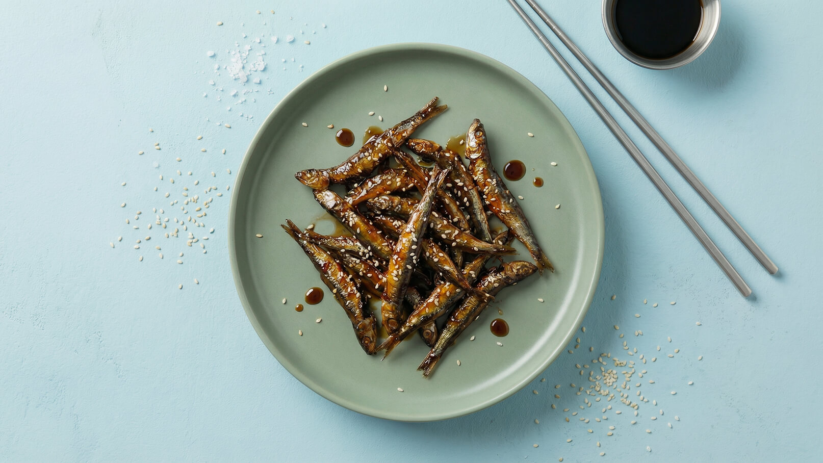 Ein Teller mit gebratenen kleinen Fischen, die mit einer glänzenden Sojasauce überzogen sind. Sie sind mit Sesam bestreut und liegen auf einem hellgrünen Teller. Daneben befinden sich Essstäbchen und einige Sesamkörner auf einem blauen Untergrund.