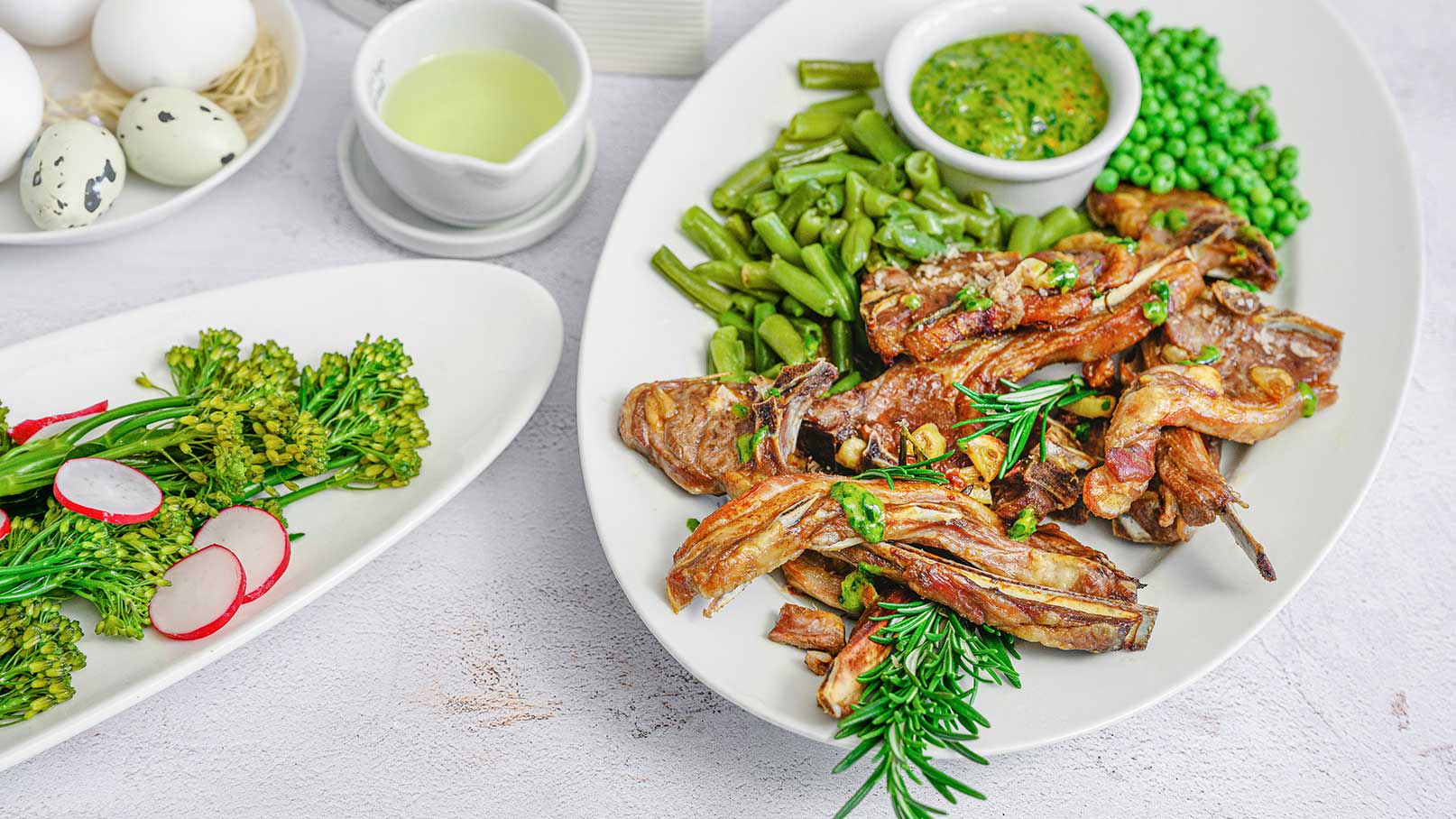 Ein Teller mit gebratenem Fleisch, garniert mit frischen Kräutern. Daneben befinden sich grüne Bohnen und Erbsen. Ein weiteres Gericht zeigt bunte Gemüsesorten, darunter Radieschen und Brokkoli. Im Hintergrund sind einige Eier und eine Schüssel mit grüner Sauce zu sehen.