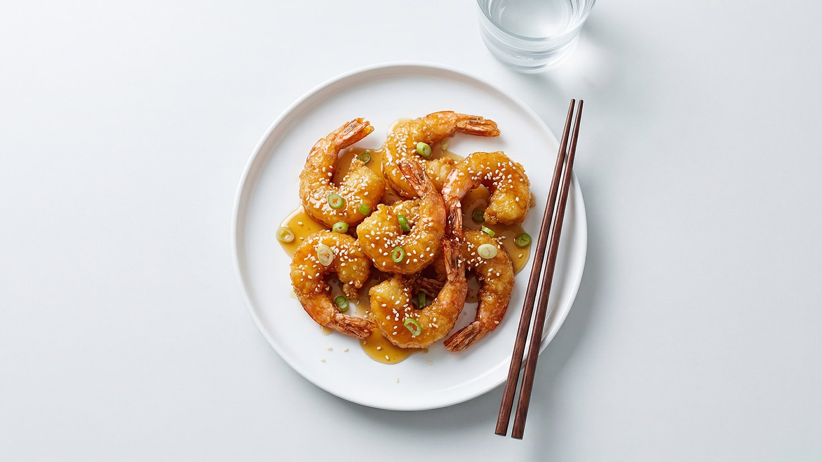 Ein Teller mit knusprigen, goldbraunen Shrimps in einer süß-sauren Sauce, garniert mit Sesam und frischen Frühlingszwiebeln. Zwei Essstäbchen liegen neben dem Teller.