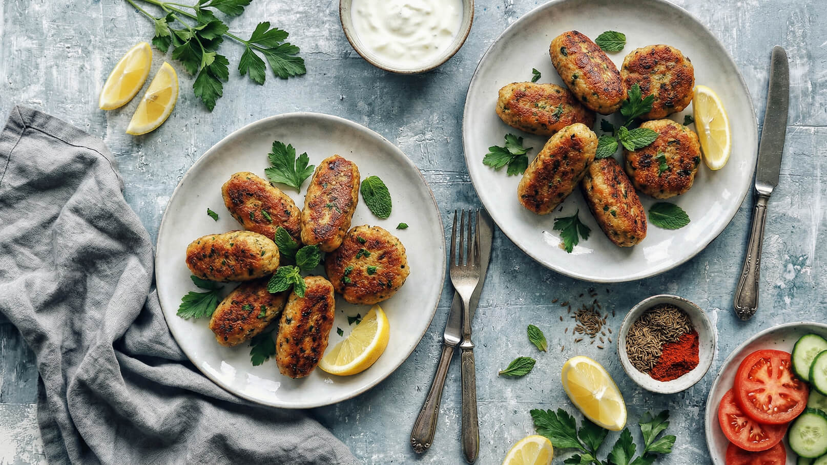 Zwei weiße Teller mit einer Vielzahl von gebratenen, goldbraunen Fleischbällchen, garniert mit frischen Kräutern. Neben den Tellern liegen Zitronenscheiben und eine kleine Schüssel mit Joghurt-Dip. Der Hintergrund ist mit grünen Kräutern und Gewürzen geschmückt.