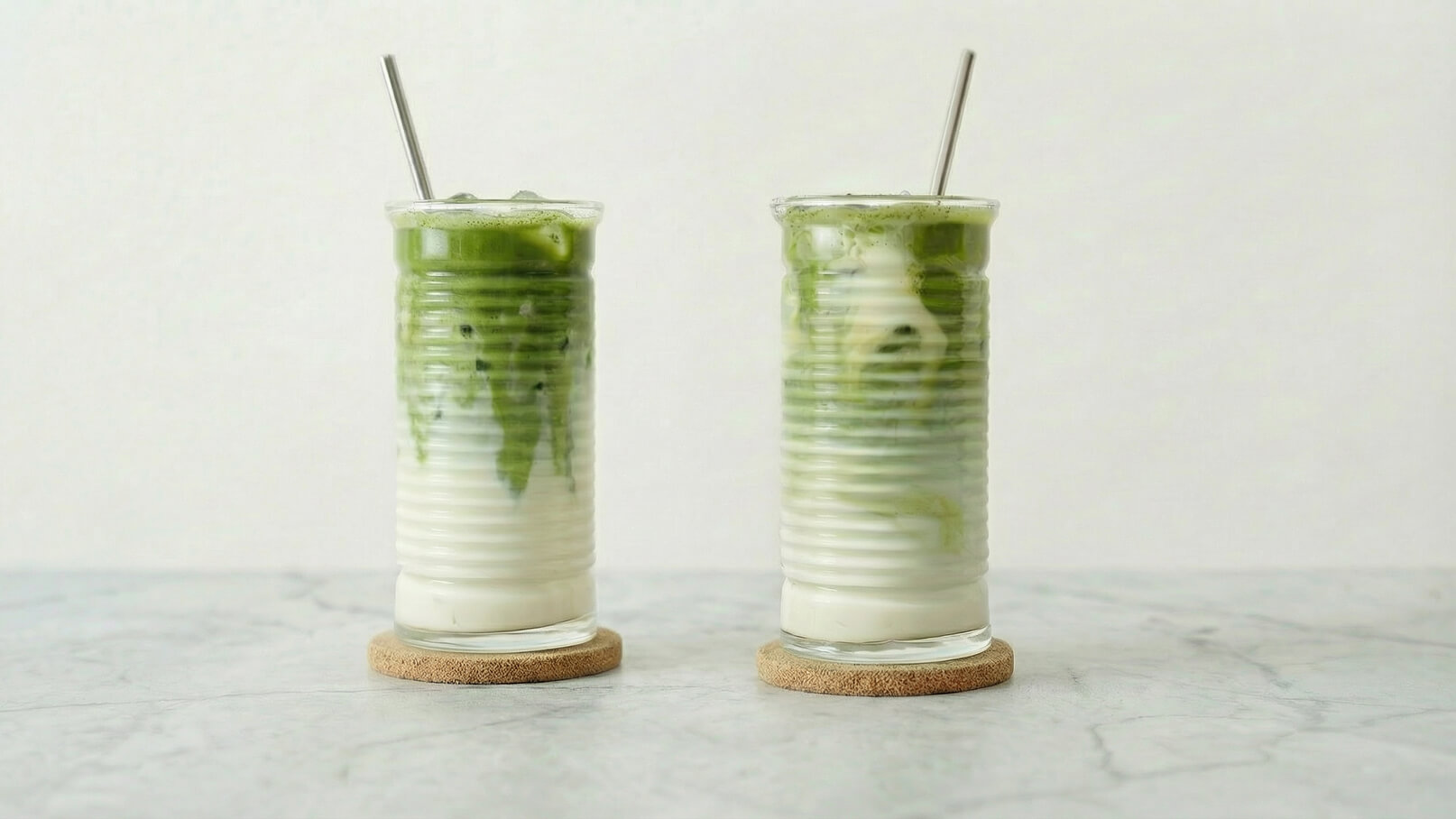 Zwei hohe Gläser mit einem geschichteten Getränk, das grüne Matcha-Schicht und eine weiße Milchschicht enthält. Beide Gläser stehen auf Korkuntersetzern auf einer hellen, marmorierten Oberfläche. Ein Strohhalm steckt in jedem Glas.