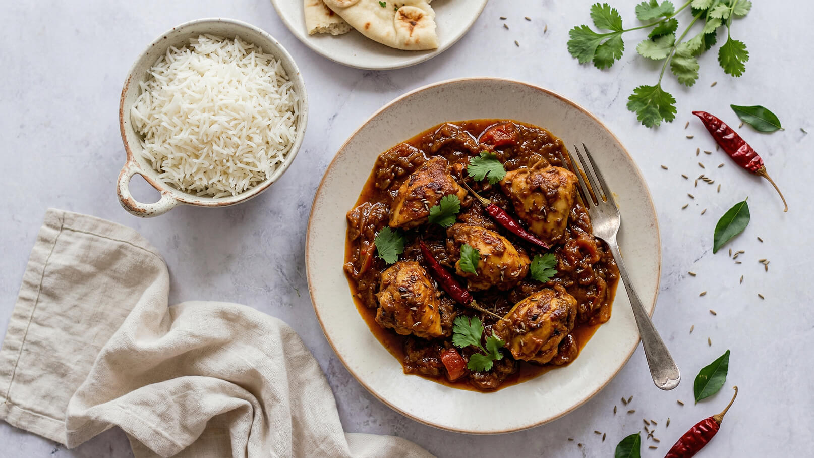 Eine Schüssel mit herzhaftem Curry, garniert mit frischen Korianderblättern und roten Chilischoten, steht auf einem Teller. Nebenan befinden sich dampfender Reis in einer kleinen Schüssel und ein Stück Naan-Brot auf einem weiteren Teller.