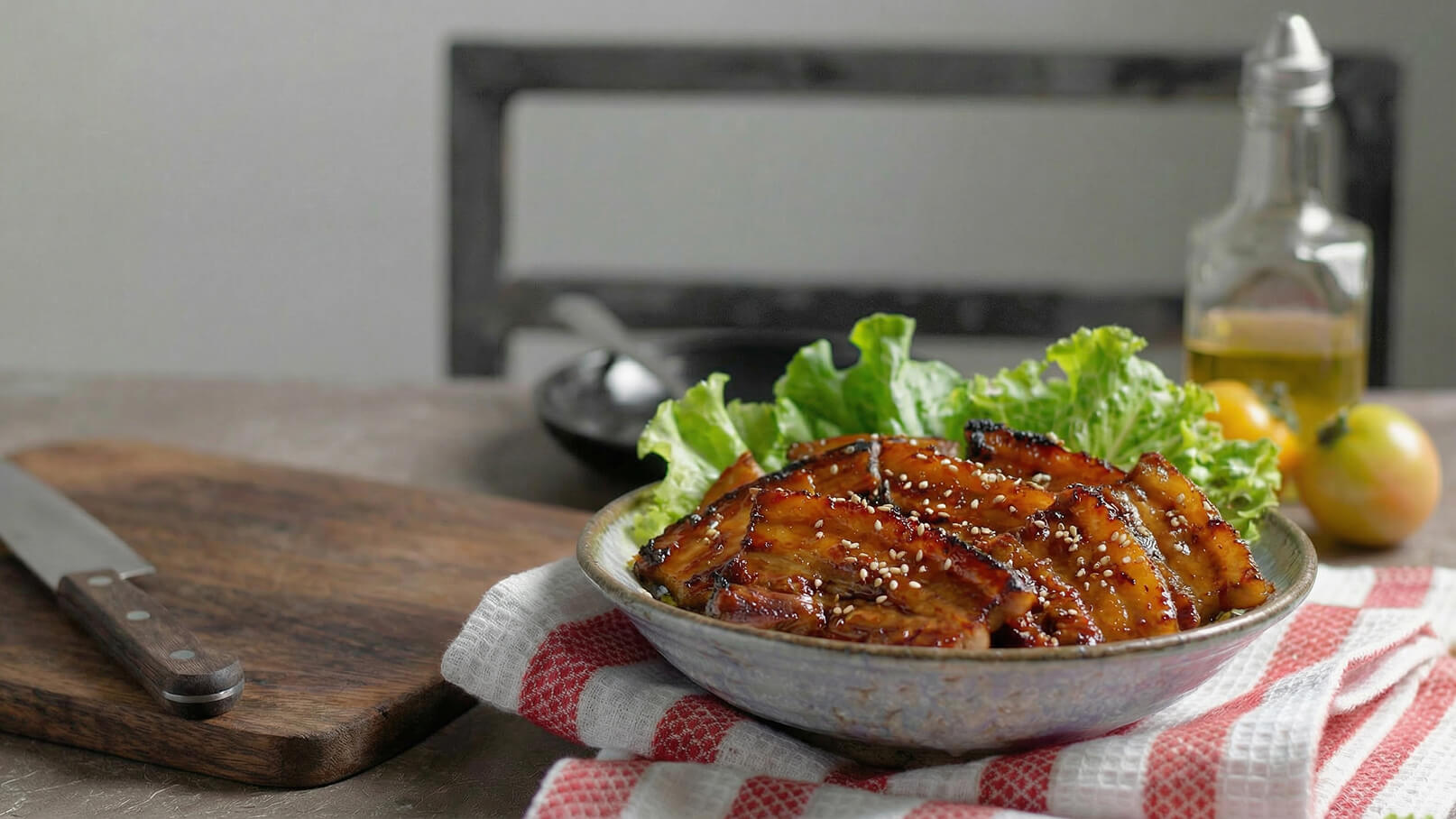 Eine Schüssel mit gegrilltem Fleisch in einer süßen Marinade, garniert mit Sesam, liegt auf einem Bett aus frischem grünen Salat. Die Schüssel steht auf einem roten und weißen karierten Küchentuch. Im Hintergrund sind eine Flasche Öl und einige neue Früchte zu sehen.