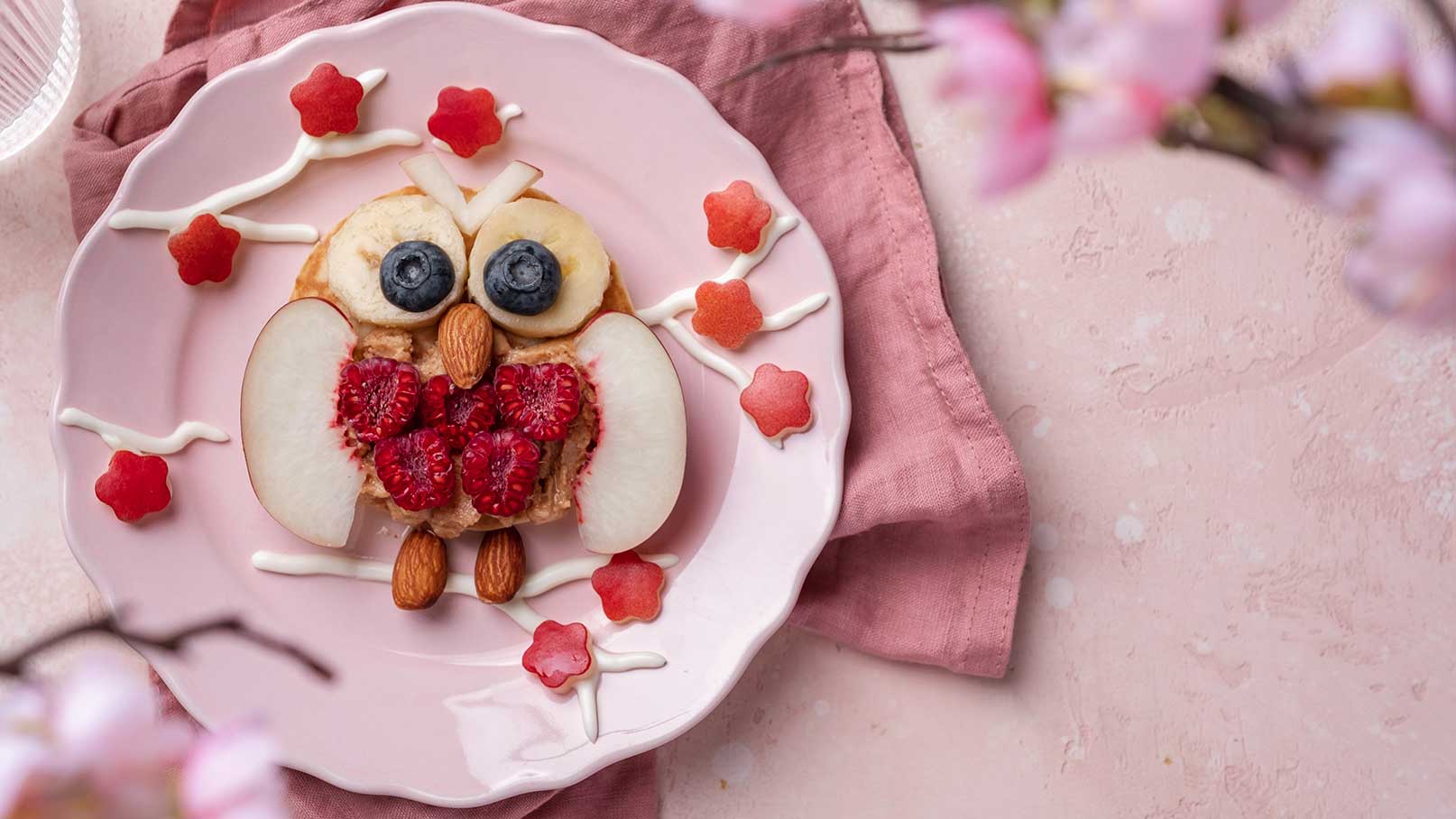 Ein Teller mit einer kunstvoll gestalteten Speise in Form einer Eule. Die Eule besteht aus Bananenscheiben, Heidelbeeren und Mandeln, während die Augen aus frischen Früchten und die Flügel aus Apfelscheiben bestehen. Um die Eule herum sind dekorative, essbare Blumen und Streifen aus Sahne.
