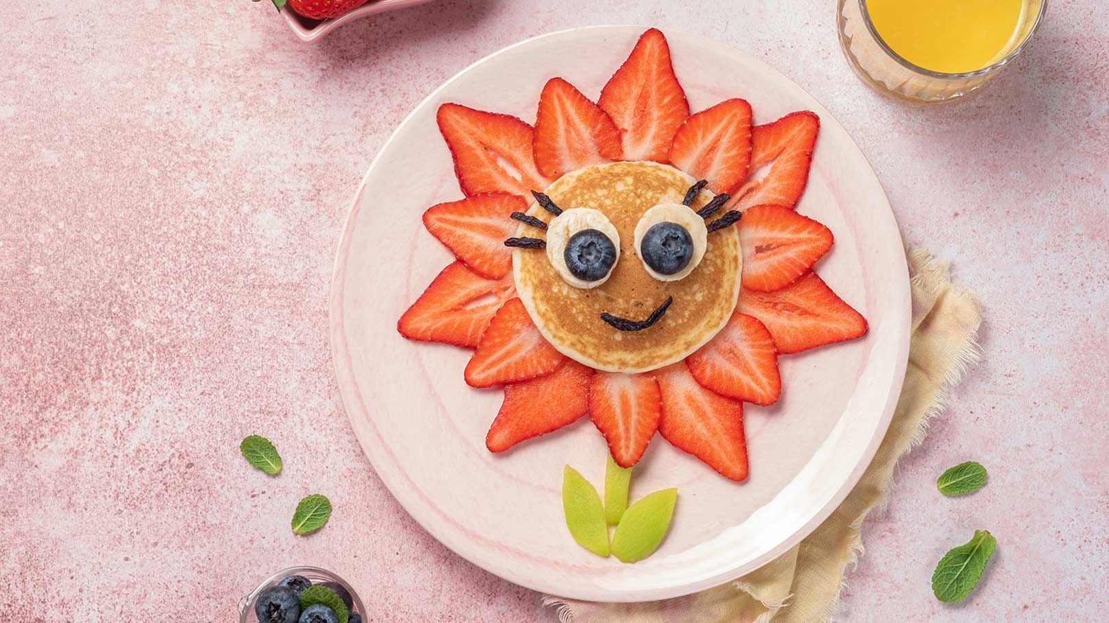 Ein Teller mit einem Pfannkuchen, der wie eine Blume dekoriert ist. Erdbeeren bilden die Blütenblätter, während der Pfannkuchen das Blütenzentrum darstellt. Augen aus Bananen und Heidelbeeren sowie ein lächelndes Gesicht. Grünes Blattwerk aus Apfelstücken ergänzt das Bild.