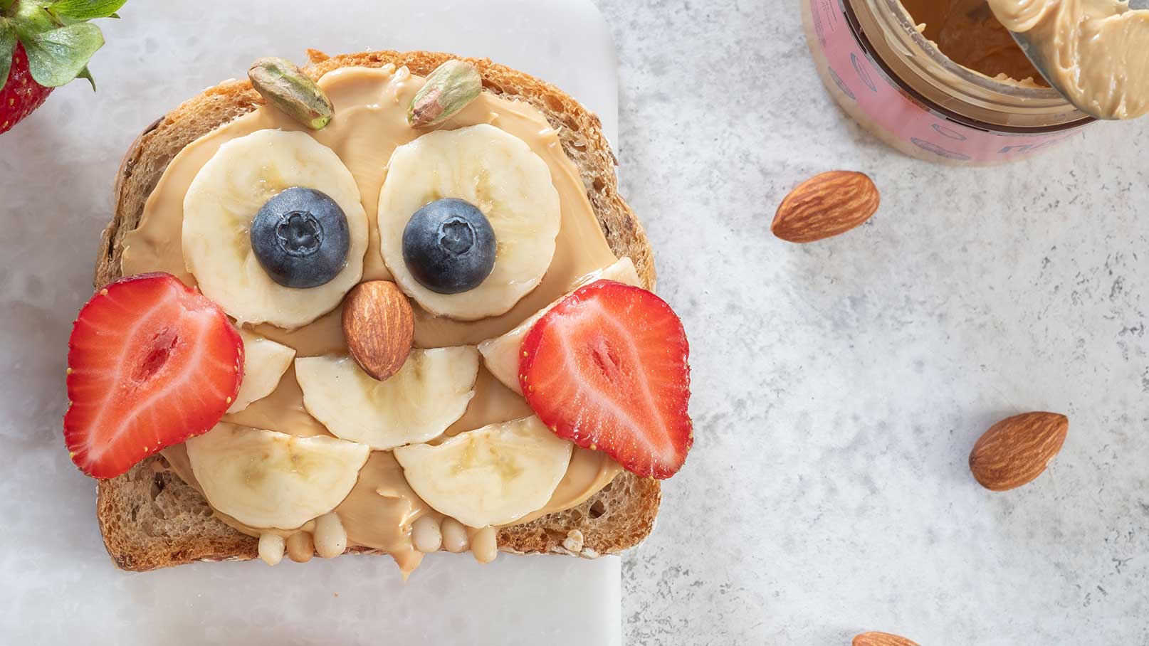 Ein Stück Vollkornbrot ist mit Erdnussbutter bestrichen und kreativ mit Bananenscheiben, einer Heidelbeere und einer Erdbeere dekoriert, um ein Gesicht einer Eule darzustellen. Mandeln bilden den Schnabel. Im Hintergrund sind einige Mandeln sichtbar.