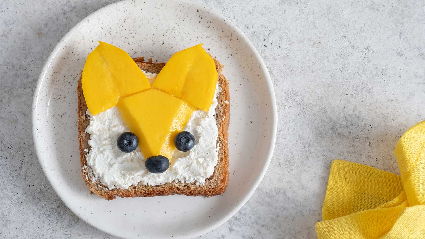 Ein Stück Vollkornbrot liegt auf einem Teller. Darauf ist eine kreative Gestaltung mit Frischkäse, Mango und Heidelbeeren, die ein Tiergesicht darstellen. Die Ohren sind aus Mangoscheiben, und die Augen sowie die Nase sind aus Heidelbeeren.