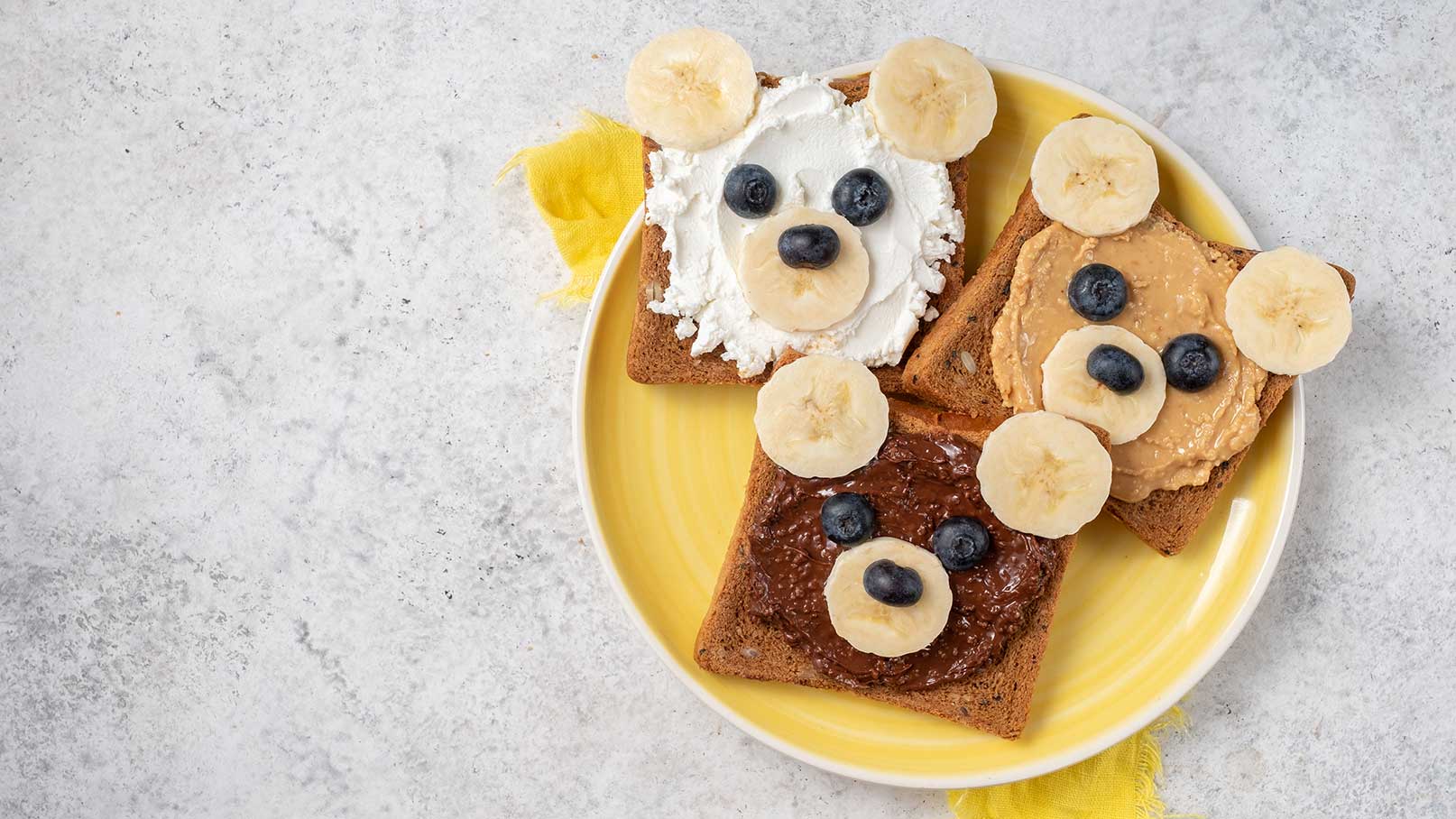 Drei mit Lebensmitteln dekorierte Brotscheiben in Form von Bärenköpfen auf einem gelben Teller. Die Brote sind mit verschiedenen Aufstrichen (frischkäse, Erdnussbutter, Schokoladencreme) belegt und mit Bananen- und Heidelbeerstücken verziert, die als Augen und Ohren dienen.