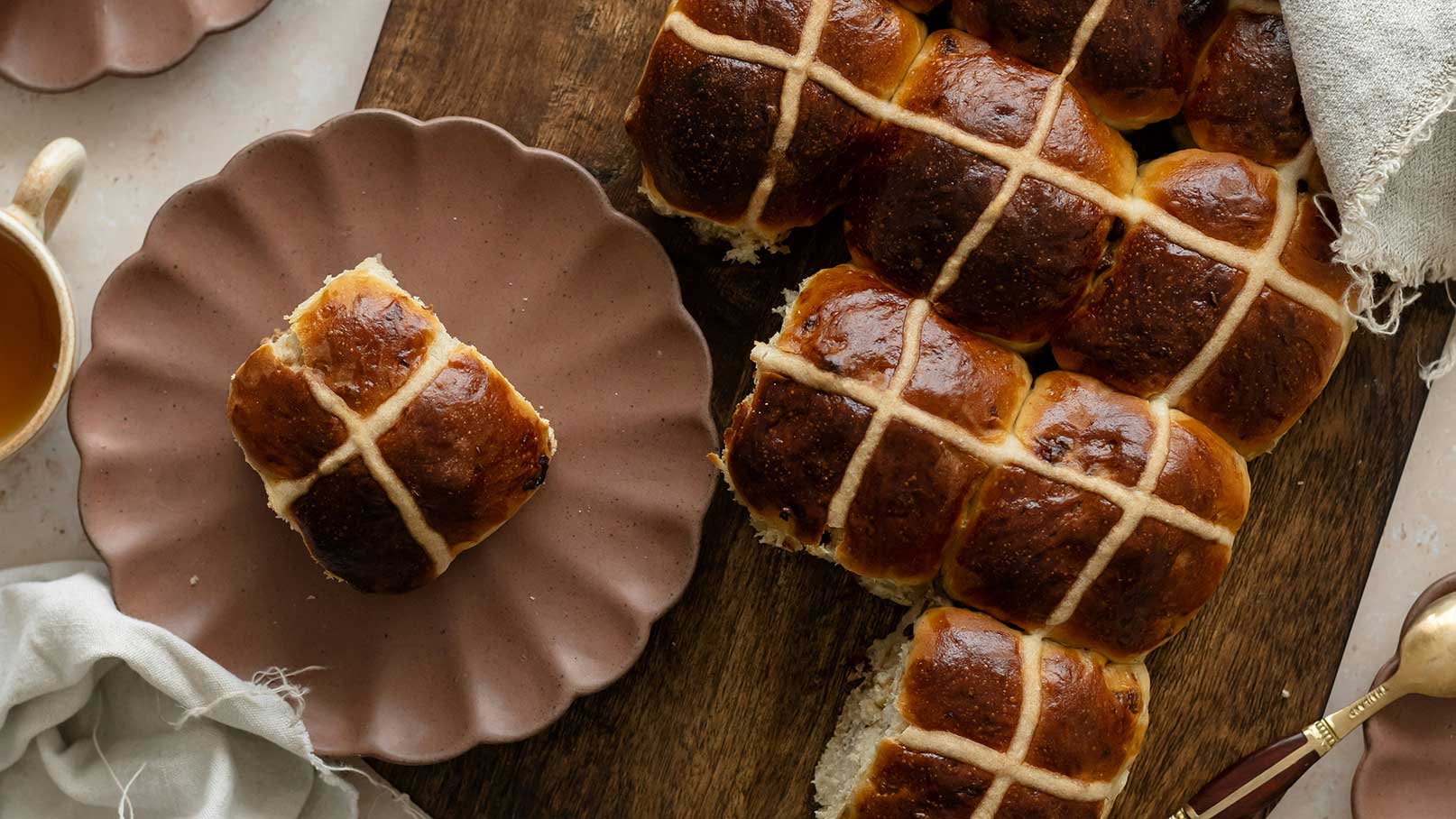 Auf dem Holzbrett liegen mehrere frisch gebackene Hot Cross Buns, ein traditionelles süßes Brötchen mit einem Kreuz aus Teig. Ein einzelnes Brötchen ist auf einem braunen Teller angerichtet, daneben steht eine Tasse Tee.