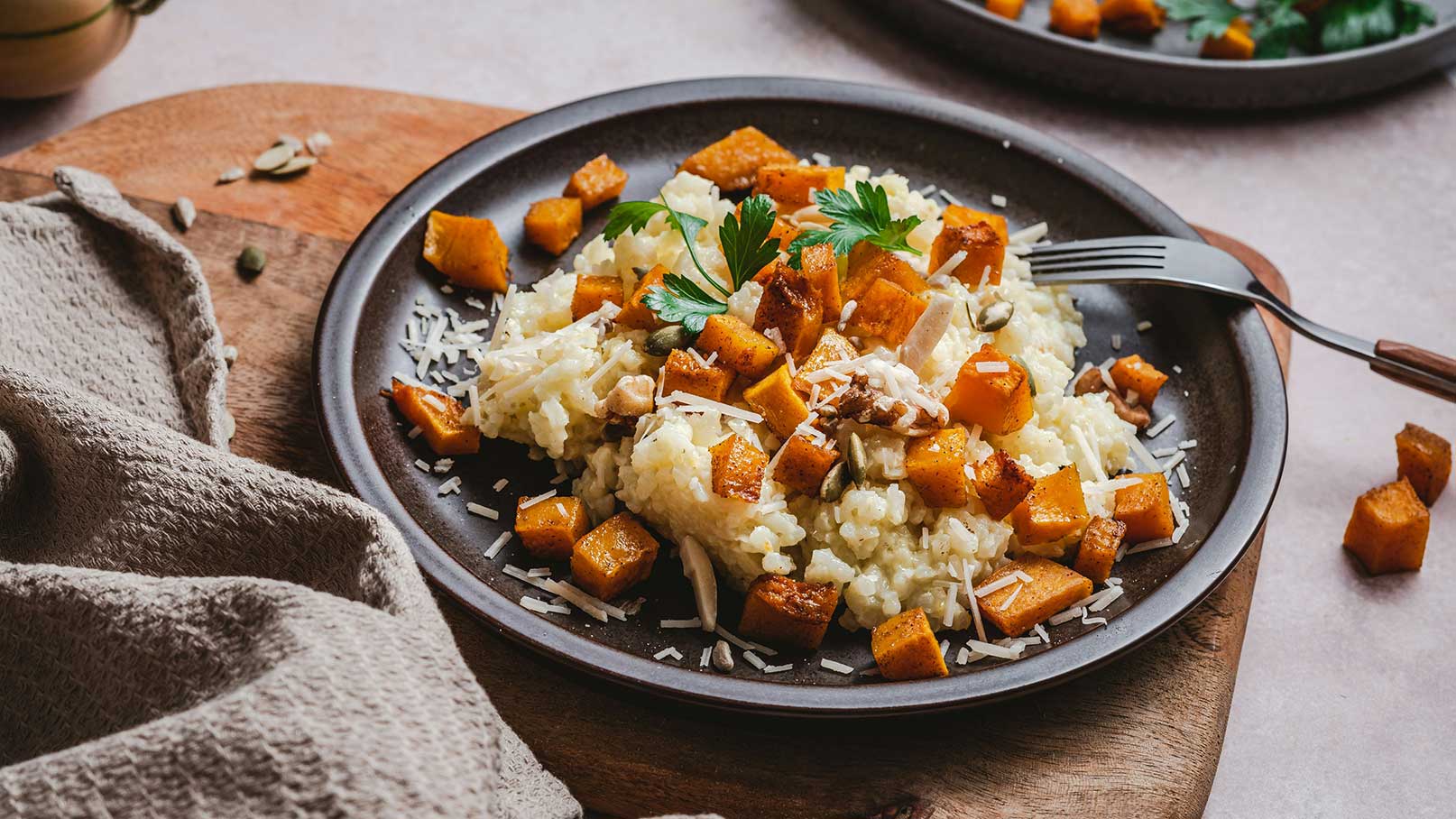 Ein Teller mit Risotto, garniert mit gebratenen Kürbiswürfeln und einem Hauch von geriebenem Käse. Frische Petersilie sorgt für einen farblichen Akzent. Im Hintergrund sind unregelmäßig geschnittene Kürbisstücke zu sehen.