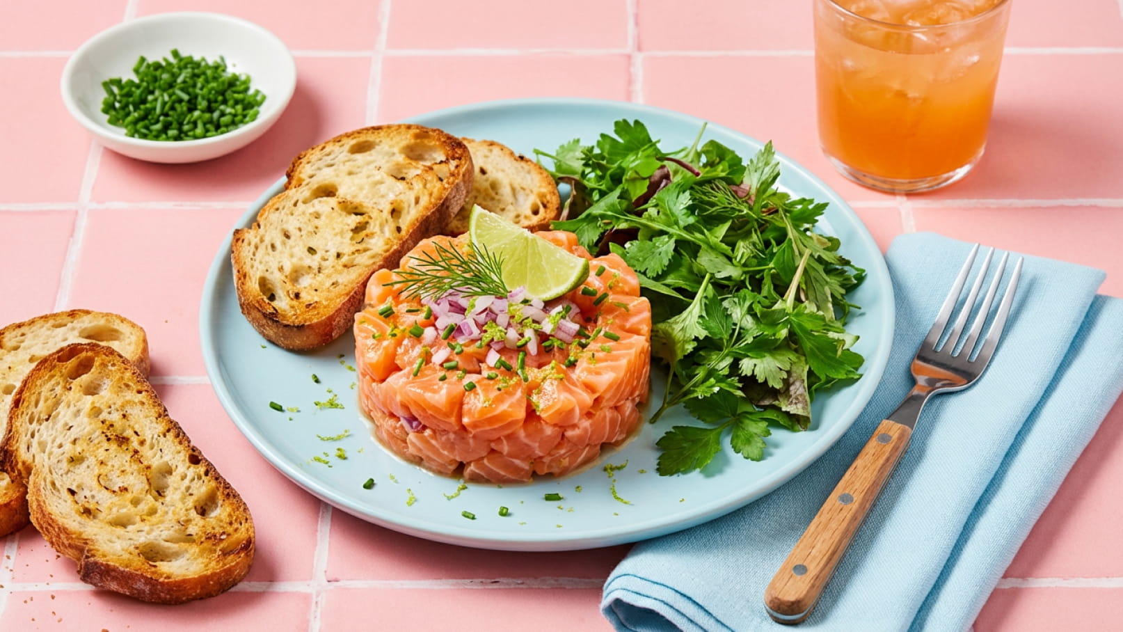 Ein Teller mit Lachs-Tatar, garniert mit Zwiebeln, Dill und Limettenscheibe, umgeben von frischem Salat und geröstetem Brot. Daneben ein Glas mit einem orangefarbenen Getränk. Der Hintergrund ist rosa.