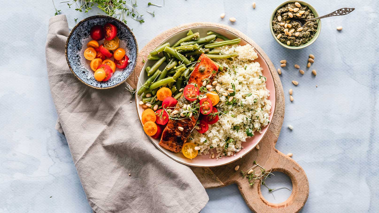 Ein Teller mit gebratenem Lachs, grünem Gemüse und Couscous, garniert mit Kirschtomaten und Kräutern. Daneben befindet sich eine Schüssel mit bunten Cherrytomaten und ein kleiner Behälter mit Pesto. Der Tisch ist mit einer geschmackvollen Tischdecke und einem Holzbrett umgeben.