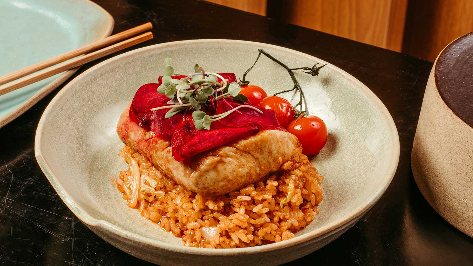 Ein Teller mit gebratenem Hähnchenfilet, serviert auf einer Schicht würzigem Reis. Darüber liegen rote Rüben und frische Kräuter. Neben dem Hähnchen sind einige Kirschtomaten platziert. Der Hintergrund ist minimalistisch gehalten, wobei der Fokus auf dem Gericht liegt.