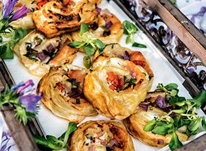 Ein rustikaler Holztablett enthält frisch gebackene, gefüllte Blätterteigrollen. Die Rollen sind mit Gemüse und Kräutern garniert und präsentieren sich appetitlich. Im Hintergrund sind dekorative Blumen sichtbar, die das Gericht umrahmen.
