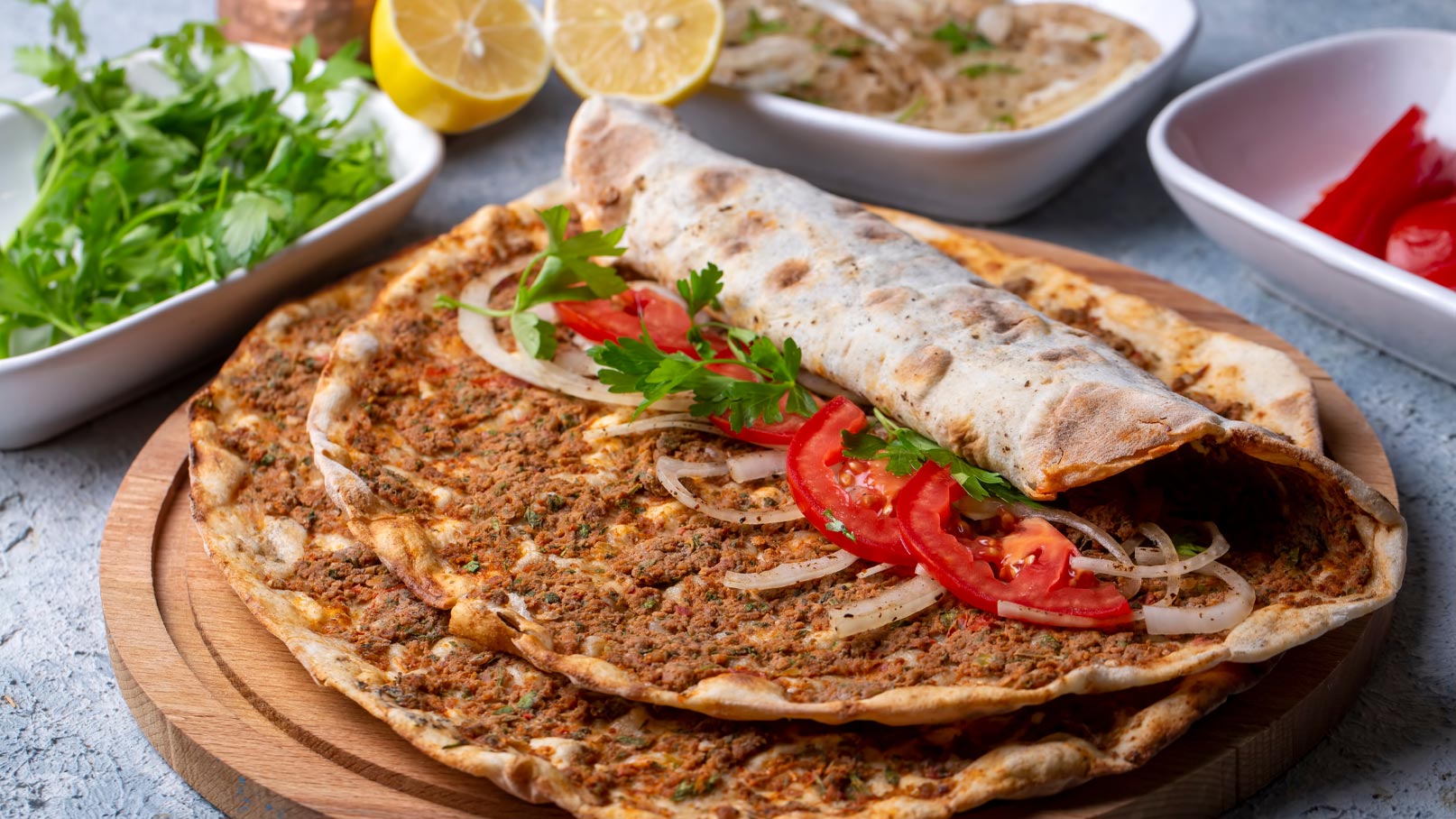 Auf einem Holzbrett liegen mehrere flache Brote mit würziger Füllung. Ein Brot ist aufgerollt und mit frischen Tomatenscheiben, Zwiebeln und Blattgrün garniert. Im Hintergrund sind Zitronen und kleine Schalen mit weiteren Zutaten sichtbar.