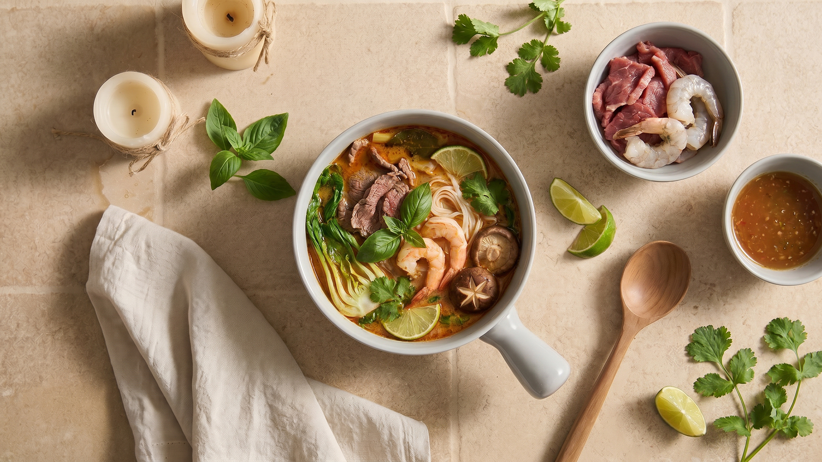 Eine große Schüssel mit Ramennudeln, Garnelen, Rindfleisch und Gemüse in einer würzigen Brühe. Frische Kräuter und Limettenscheiben dekorieren das Gericht. Neben der Schüssel befinden sich kleine Schalen mit rohem Fleisch und einer Sauce sowie eine Holzlöffel und eine hellbeige Serviette.