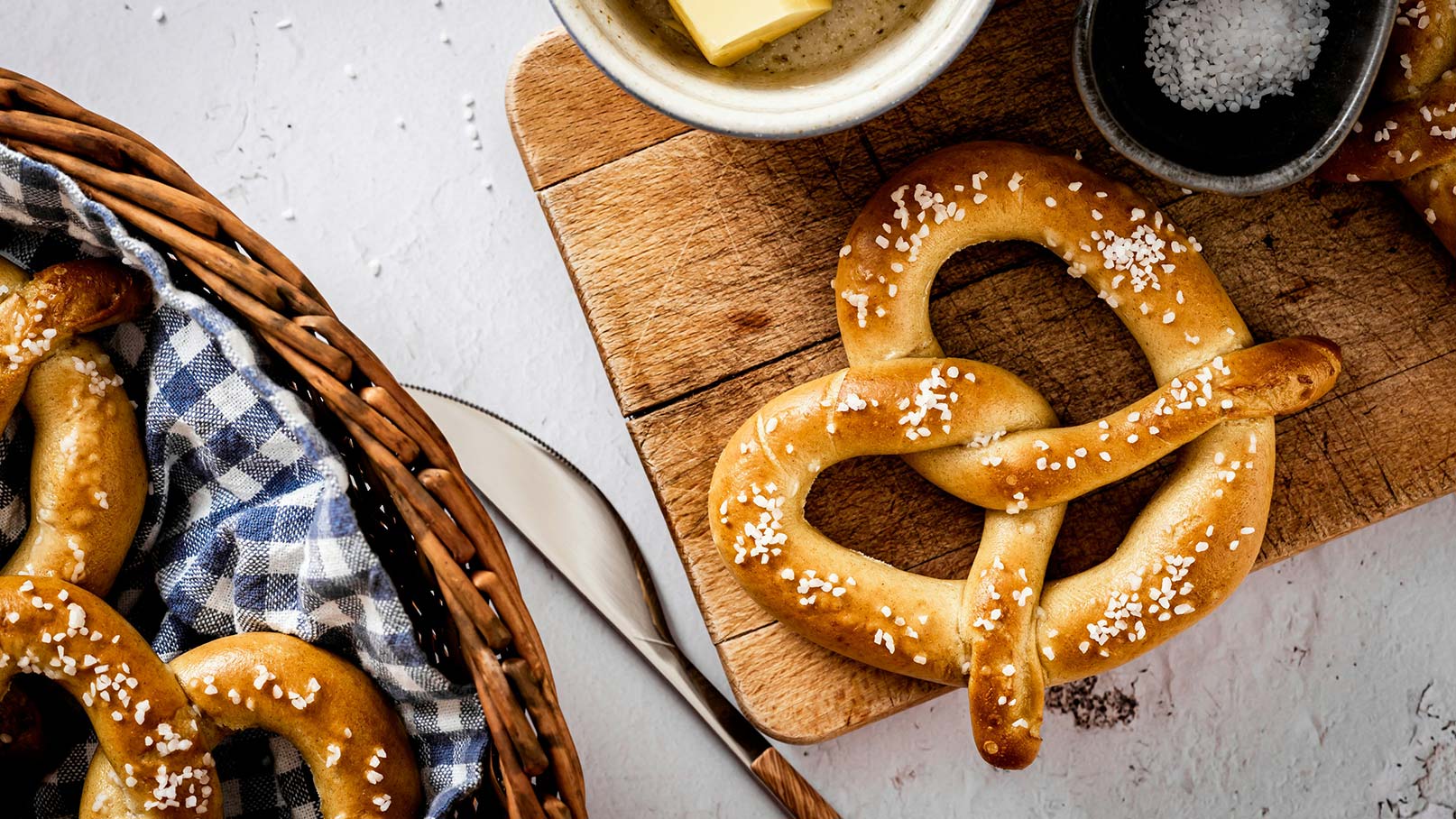 Ein geflochtenes Brezel auf einem Holzbrett, verziert mit grobem Salz. Daneben befindet sich ein kleiner Teller mit Butter und ein weiterer mit Salz. Im Hintergrund sieht man einen Korb mit zusätzlichen Brezeln, eingewickelt in ein kariertes Tuch.