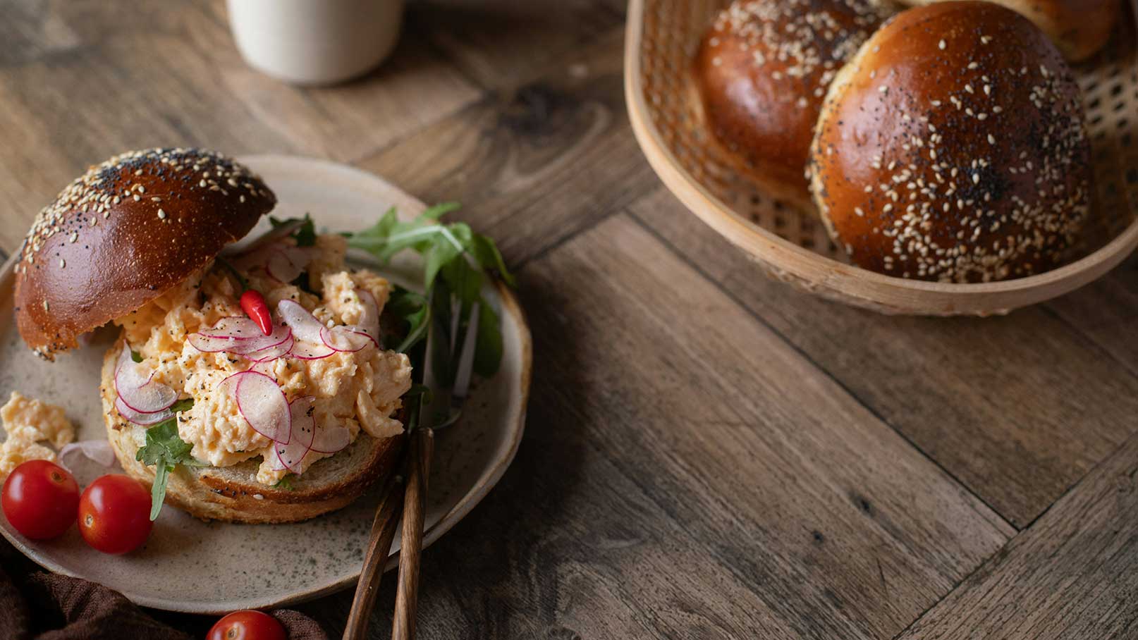 Ein rustikaler Brotburger ist mit einer cremigen Füllung, beigemischt mit Rucola und Radieschenscheiben, belegt. Eine kleine rote Chilischote sorgt für einen Farbtupfer. Daneben liegen einige Kirschtomaten und im Hintergrund ist ein Korb mit weiteren Brötchen sichtbar.