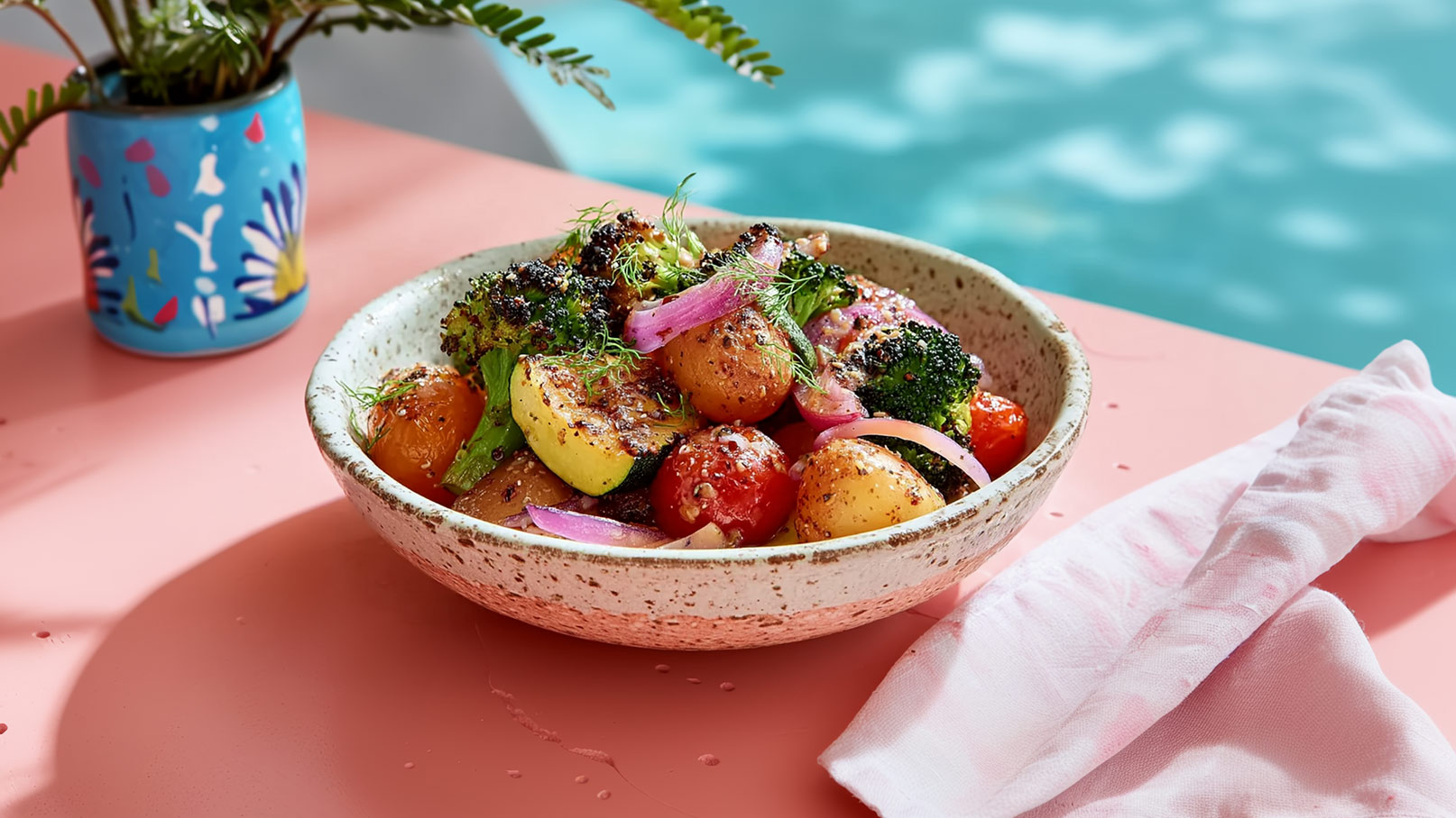 Eine Schüssel mit buntem mediterranen Gemüse ist zu sehen, darunter Kartoffeln, Brokkoli, Kirschtomaten und rote Zwiebeln, die gebraten und gewürzt sind. Die Schüssel steht auf einem rosa Tisch, neben ihr liegt ein pinkes Tuch.