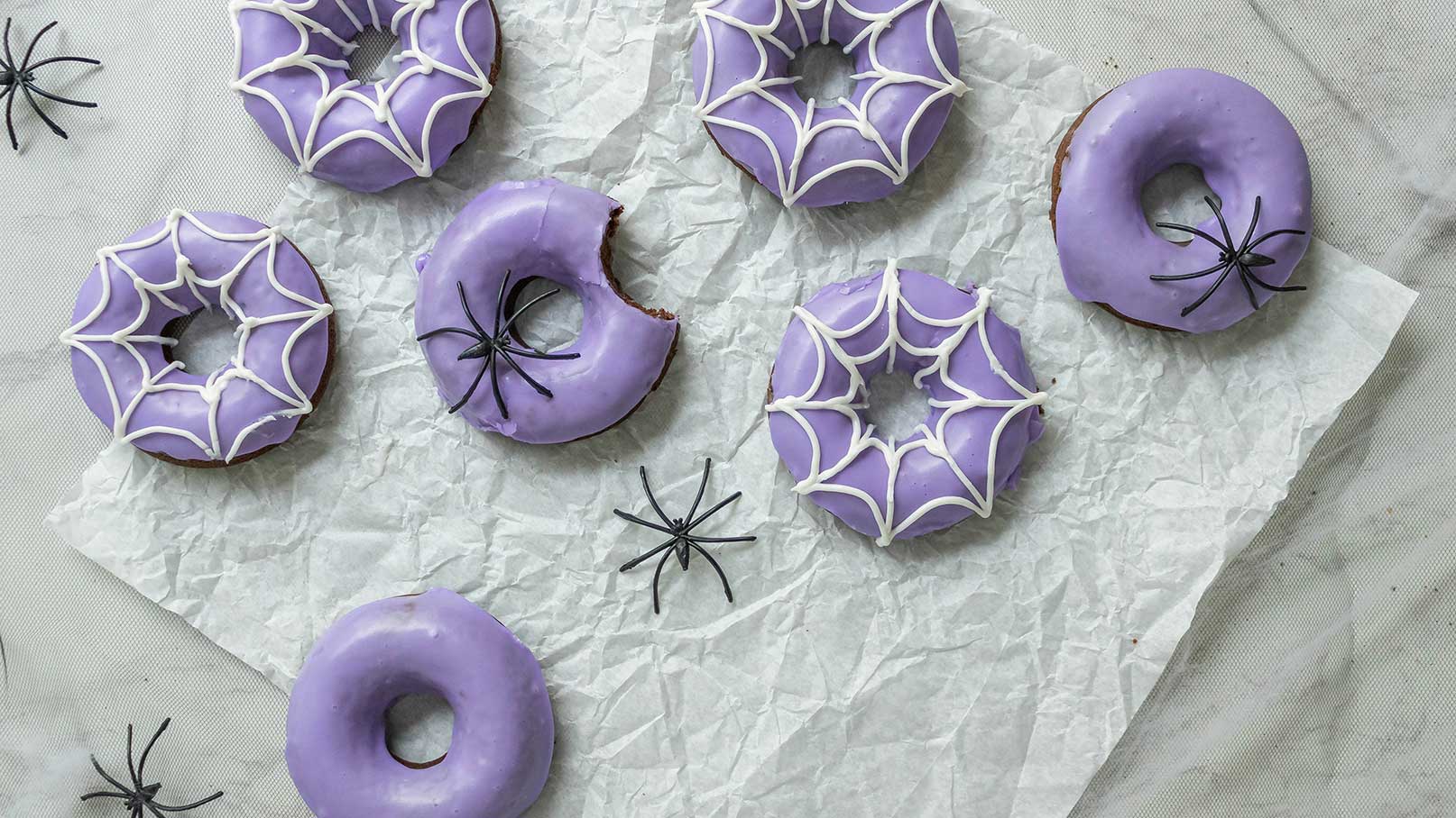 Sechs lila Donuts liegen auf zerknülltem Papier, einige mit einem weißen Spinnennetzmuster dekoriert. Ein Donut hat einen Biss, während schwarze Plastikspinnen daneben platziert sind. Die Anordnung ist spielerisch und thematisch an Halloween angelehnt.
