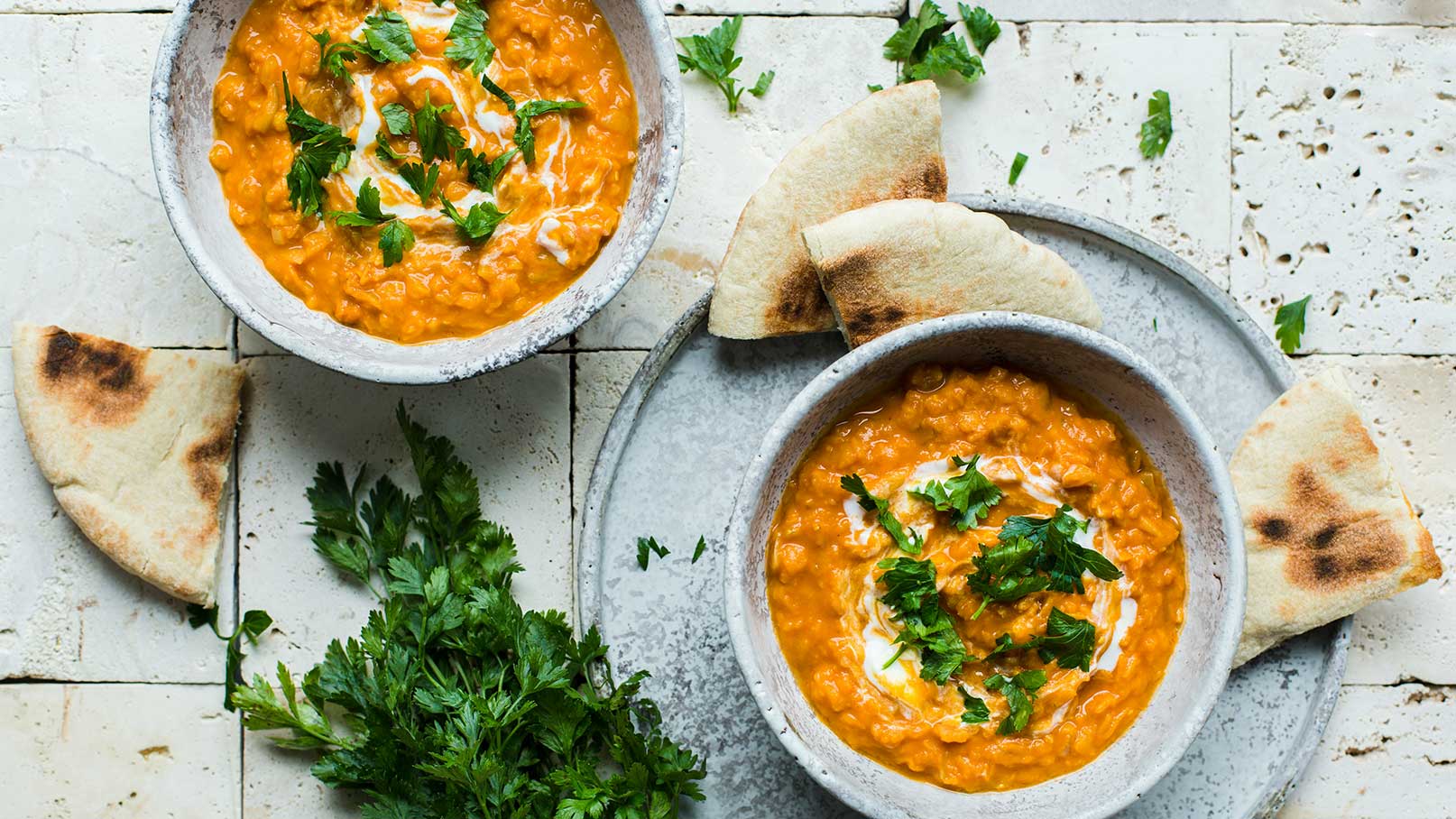 Zwei Schalen mit orangefarbener Linsensuppe, garniert mit frischem Koriander und einem Klecks Joghurt. Neben den Schalen liegen Stücke von flachem Brot auf einem grauen Teller, umgeben von frischen Korianderblättern auf einem hellen Untergrund.