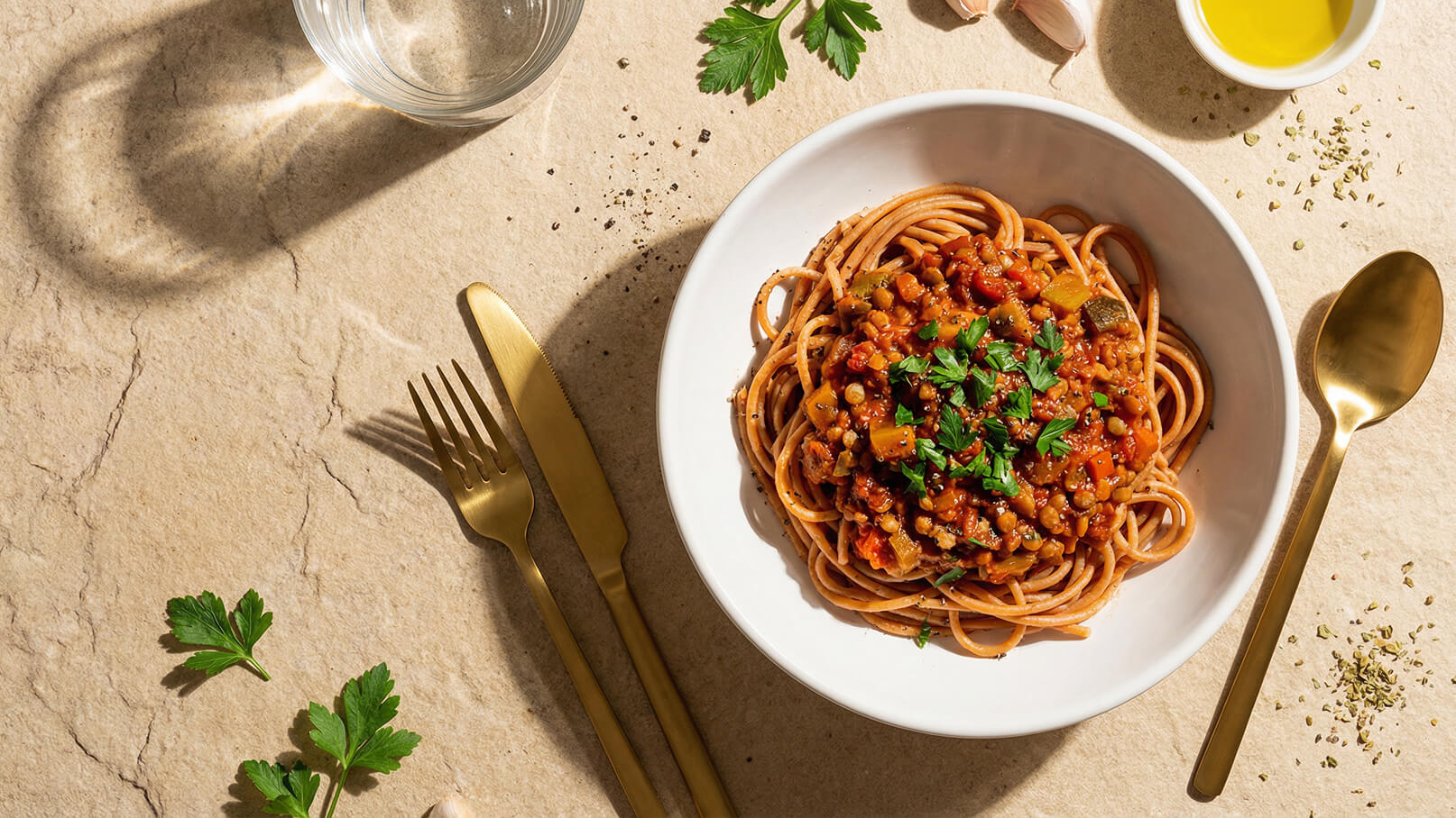 Ein Teller mit Spaghetti in einer Tomatensauce, garniert mit frischer Petersilie. Daneben liegen ein goldenes Besteckset, ein Glas Wasser und einige Küchenzutaten wie Knoblauch und Olivenöl auf einer hellen Unterlage.