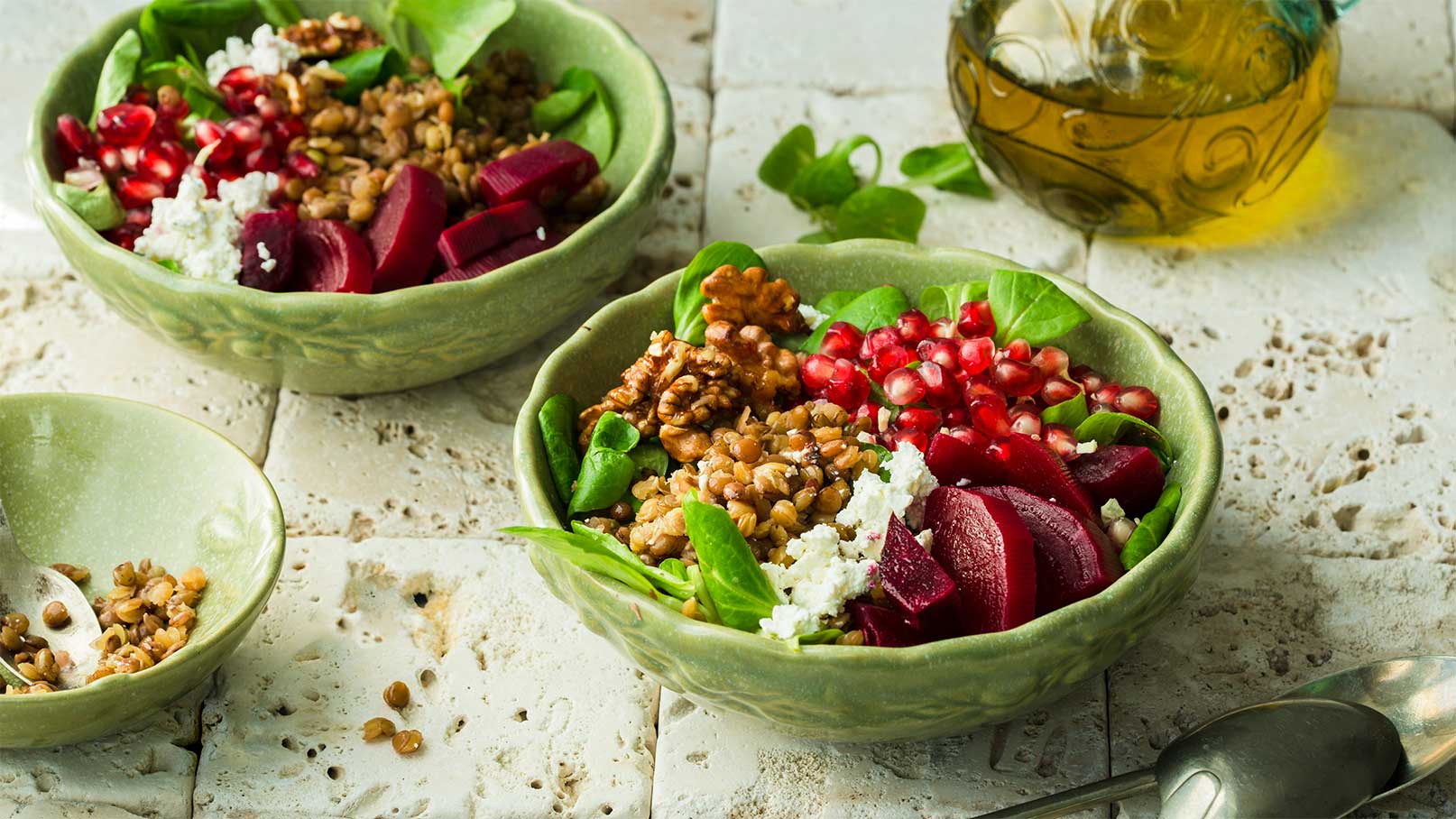 Zwei Schalen mit buntem Salat stehen auf einem hellen Untergrund. Die Salate enthalten frische Blätter, Rote Bete, Granatapfelkerne, Walnüsse und Quark. Dazu liegt eine kleine Schale mit gekochtem Weizen und ein Löffel. Im Hintergrund ist eine Glasflasche mit Öl zu sehen.