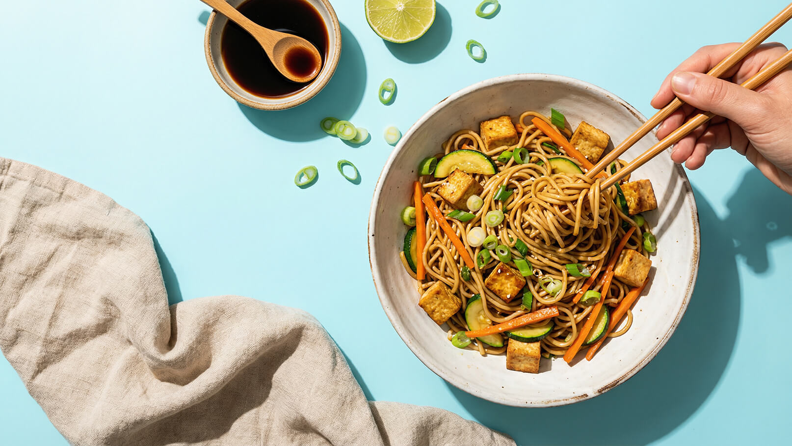Eine Schüssel mit Nudeln, Tofu und Gemüse wie Karotten und Gurken. Eine Hand hält Essstäbchen, um die Nudeln zu heben. Daneben befinden sich eine kleine Schüssel mit Sojasauce und eine Limette. Der Hintergrund ist hellblau.