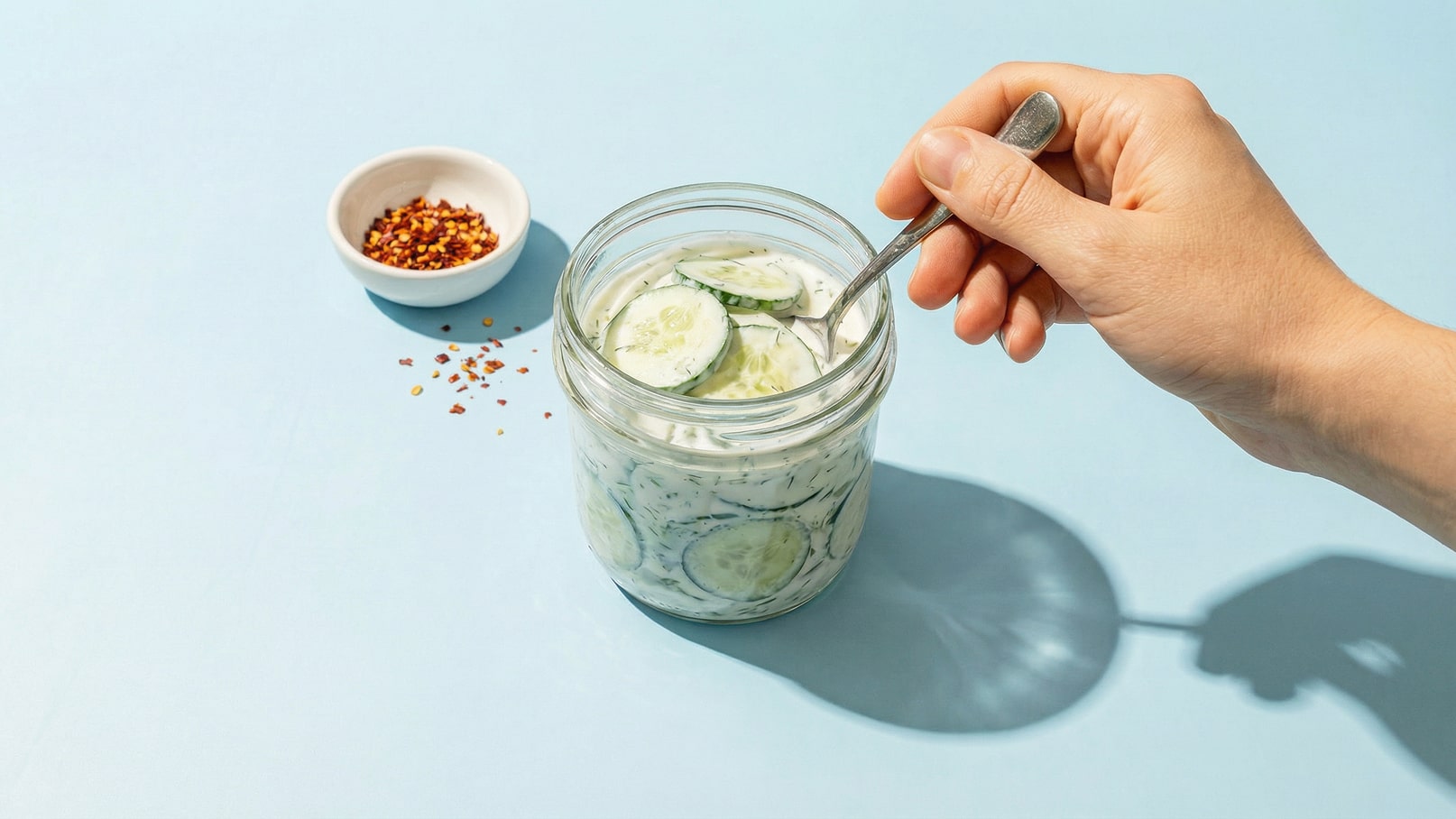 Ein Glas mit Gurkenscheiben in einer cremigen Marinade wird von einer Hand mit einem Löffel gehalten. Daneben steht eine kleine Schüssel mit roten Pfefferflocken. Der Hintergrund ist hellblau.