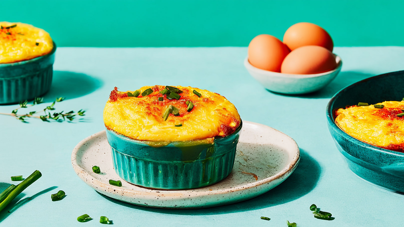 Auf einem hellblauen Tisch stehen zwei auflaufartige Gerichte in grünen Schalen, garniert mit gehacktem Schnittlauch. Daneben sind drei frische Eier in einer beige Schüssel platziert. Die Gerichte wirken appetitlich und enthalten vermutlich Eier.