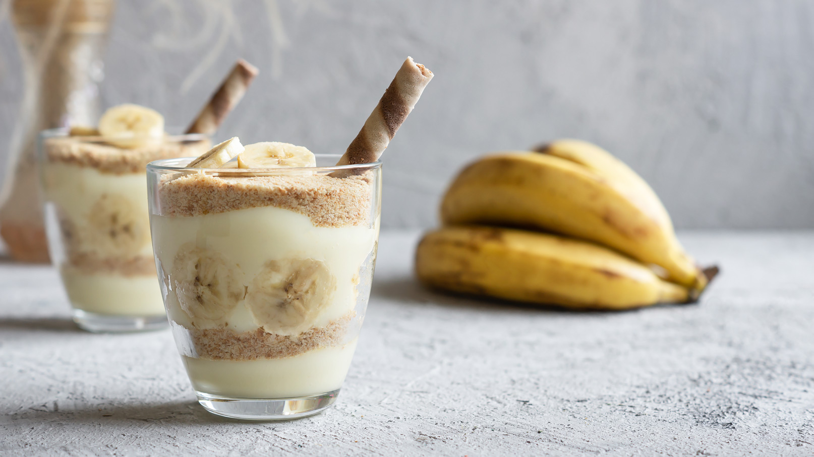 Zwei Gläser mit einem Schichtdessert aus Joghurt, zerbröselten Keksen und Bananenscheiben. Oben sind Bananenscheiben sichtbar, und ein dekorativer Waffelstift ragt aus einem der Gläser. Im Hintergrund liegen mehrere Bananen.
