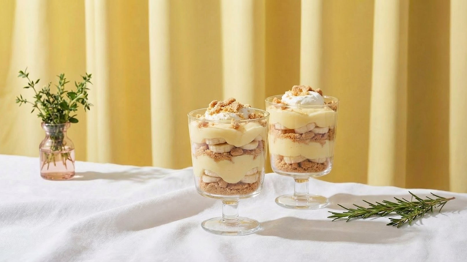 Zwei dekorative Gläser mit Schichtung aus Pudding, Bananen und zerbröselten Keksen stehen auf einem Tisch. Oben sind die Gläser mit Sahne und einem Hauch von Keksbröseln garniert. Im Hintergrund ist ein gelbes Vorhang und eine kleine Blume in einer Vase zu sehen.