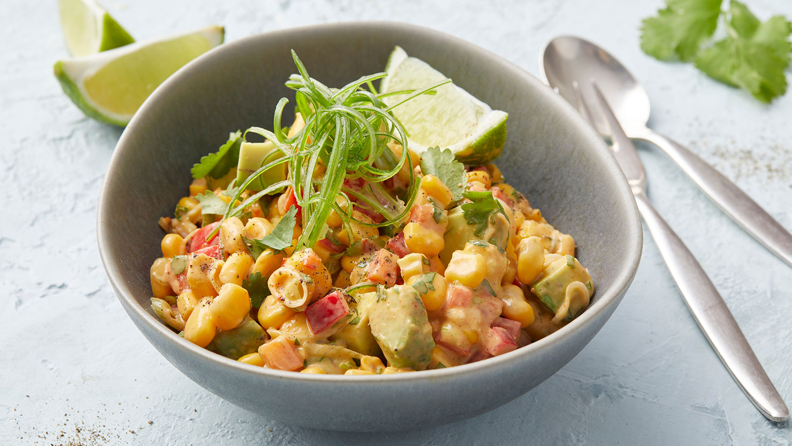 Eine Schüssel mit buntem Salat, bestehend aus Mais, Avocado, Paprika und frischen Kräutern. Der Salat ist mit einer cremigen Sauce zubereitet und mit grünen Zwiebeln garniert. Neben der Schüssel liegt eine Limettenscheibe. Der Hintergrund ist hellblau.