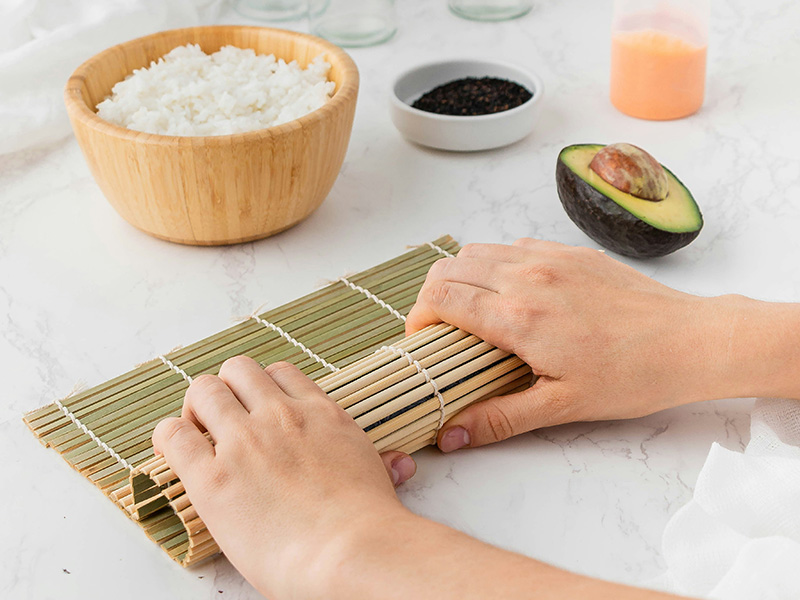Eine Hand rollt eine Sushi-Matte mit einer Füllung, die Reis und Avocado enthält. Im Hintergrund befindet sich eine Schüssel mit gekochtem Reis und kleine Schalen mit schwarzen Samen und einer rosa Sauce. Die Szene vermittelt das Thema der Sushi-Zubereitung.