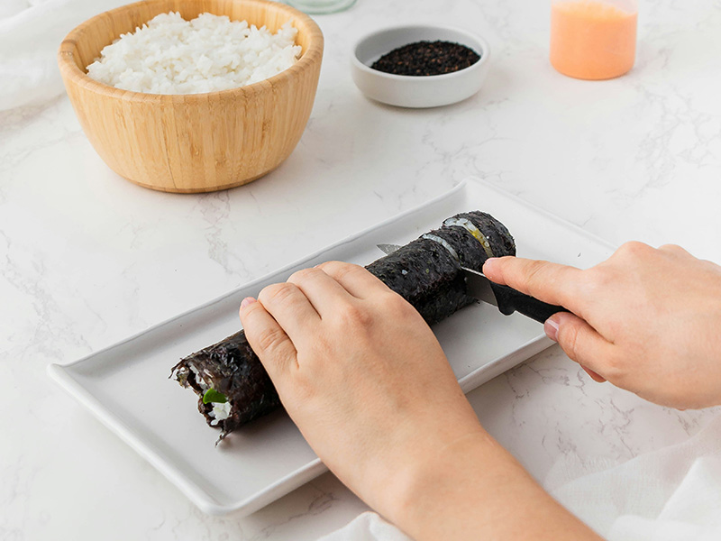 Eine Hand schneidet eine Nori-Rolle auf einem weißen Teller. Im Hintergrund steht eine Schüssel mit gekochtem Reis und eine weitere Schüssel mit schwarzem Sesam. Der Fokus liegt auf dem Schneiden der Sushi-Rolle.