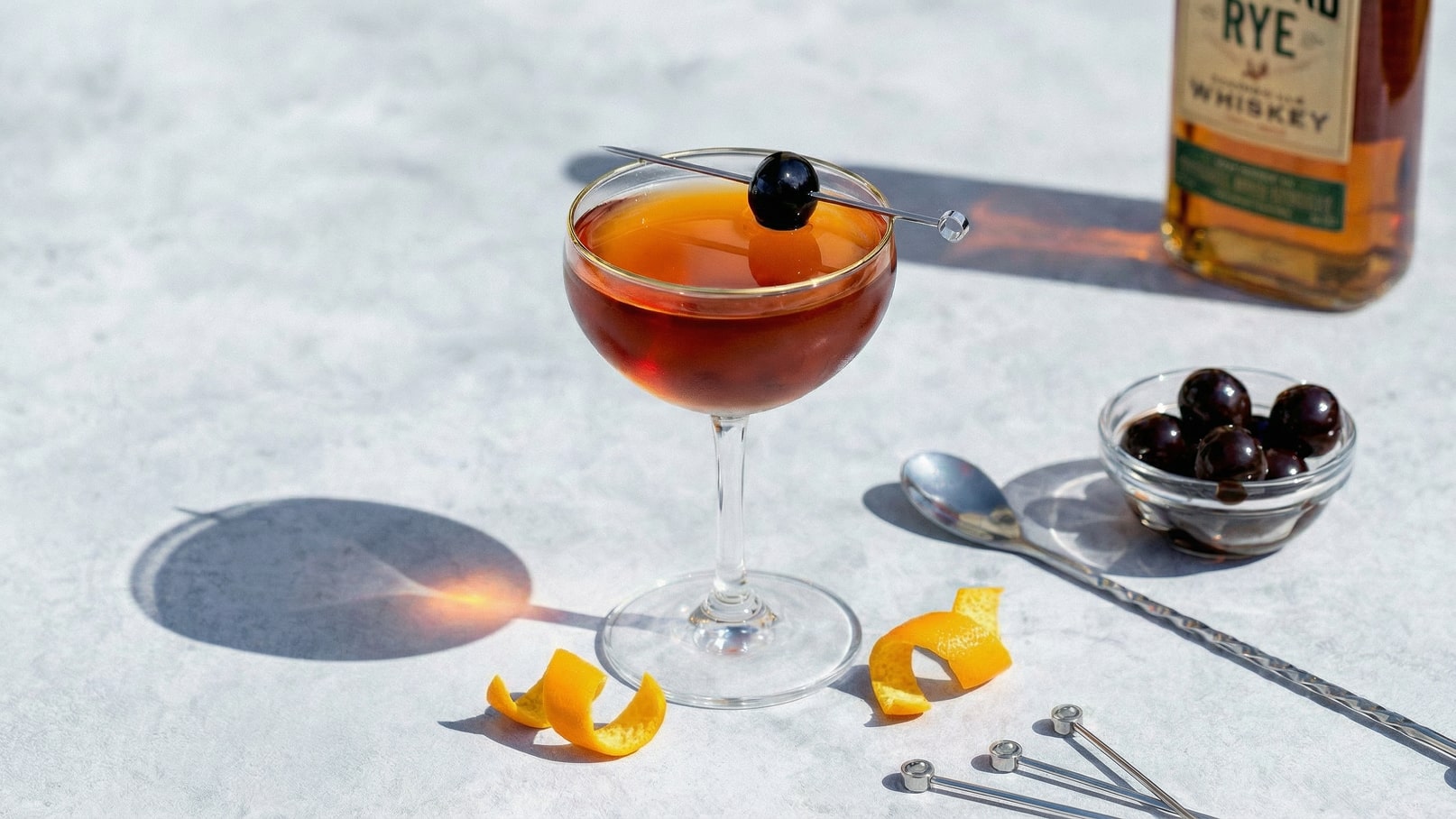 Ein elegantes Cocktailglas mit einem rötlichen Getränk, garniert mit einer Olive auf einem Zahnstocher. Daneben liegen Orangenschalen und eine Schale mit dunklen, runden Leckereien. Im Hintergrund steht eine Flasche Whiskey. Der Untergrund ist hell und strukturiert.