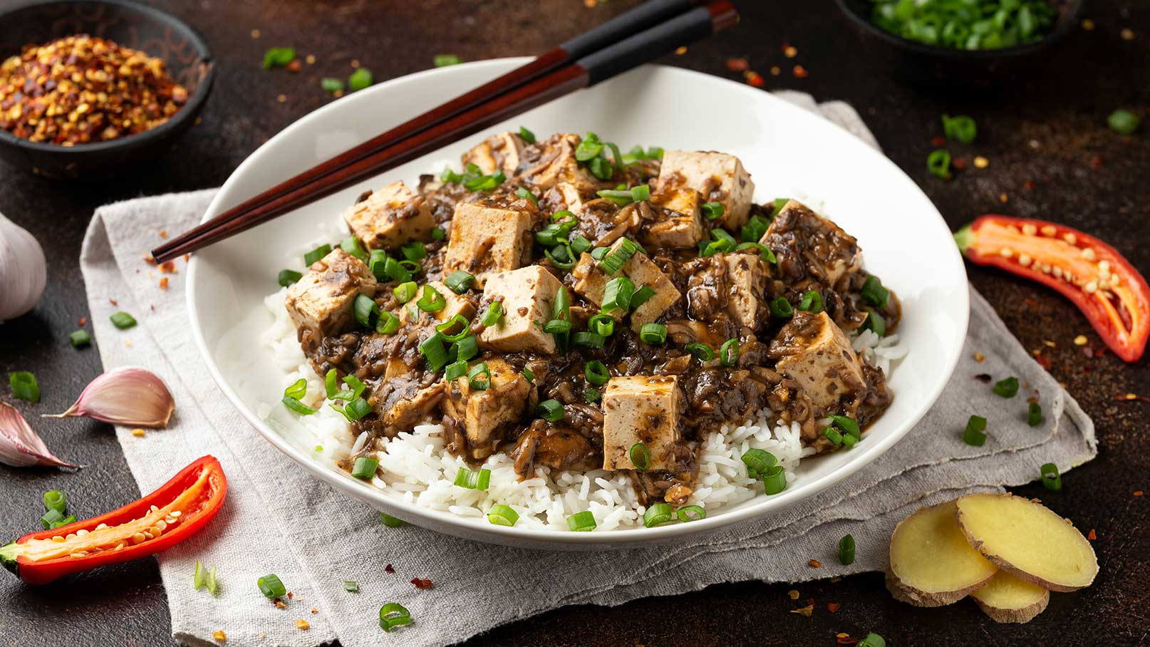 Eine Schüssel Reis mit Tofu in einer dunklen Sauce, garniert mit gehackten Frühlingszwiebeln. Daneben liegen frische Ingwerstücke und eine halbierte rote Chili. Essstäbchen sind auf der Schüssel platziert.