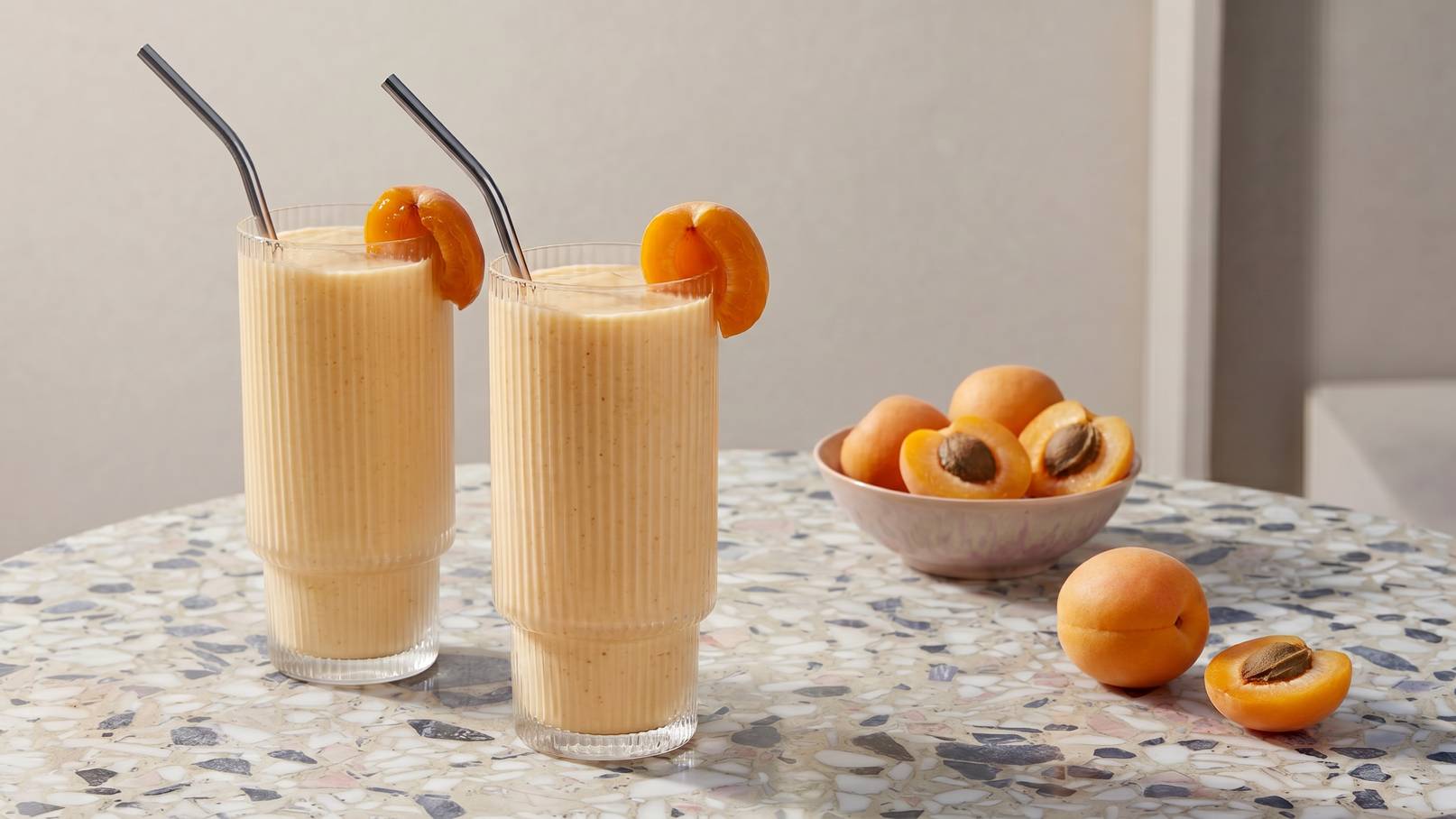 Zwei hohe Gläser mit cremigem Aprikosensmoothie stehen auf einem Tisch, jeweils mit einem Strohhalm und einer Scheibe Aprikose dekoriert. Im Hintergrund befindet sich eine Schüssel mit frischen Aprikosen.
