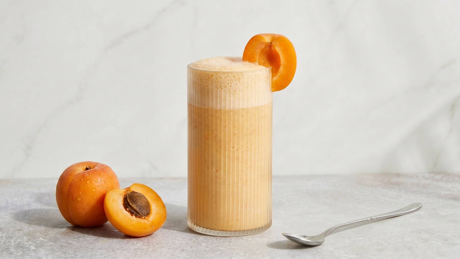Ein Glas mit einem cremigen Smoothie steht auf einer Tischplatte. Neben dem Glas liegen zwei Aprikosen, eine davon ist halbiert. Der Smoothie hat eine hellbraune Farbe und ist schaumig. Ein Löffel aus Edelstahl ist ebenfalls sichtbar.