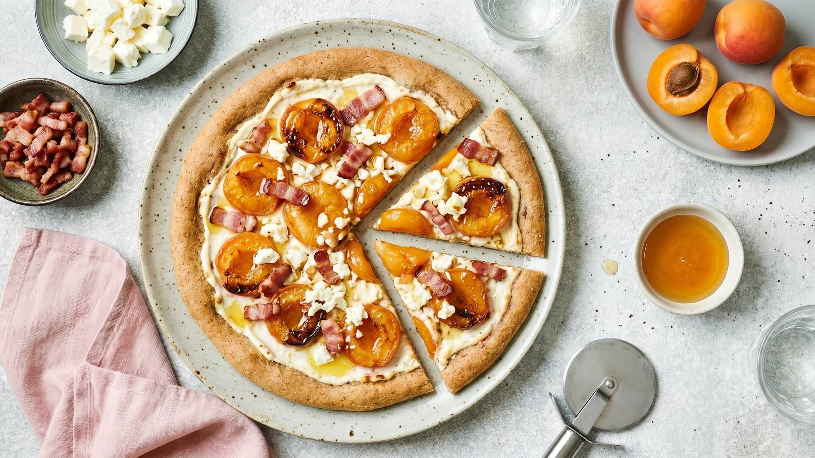 Eine Runde Pizza mit einer knusprigen Kruste, belegt mit frischen Aprikosen, Schinkenstücken und einer cremigen Füllung. Ein Stück ist bereits herausgeschnitten. Daneben sind Zutaten wie Schinken, Frischkäse und Aprikosen in Schalen platziert.