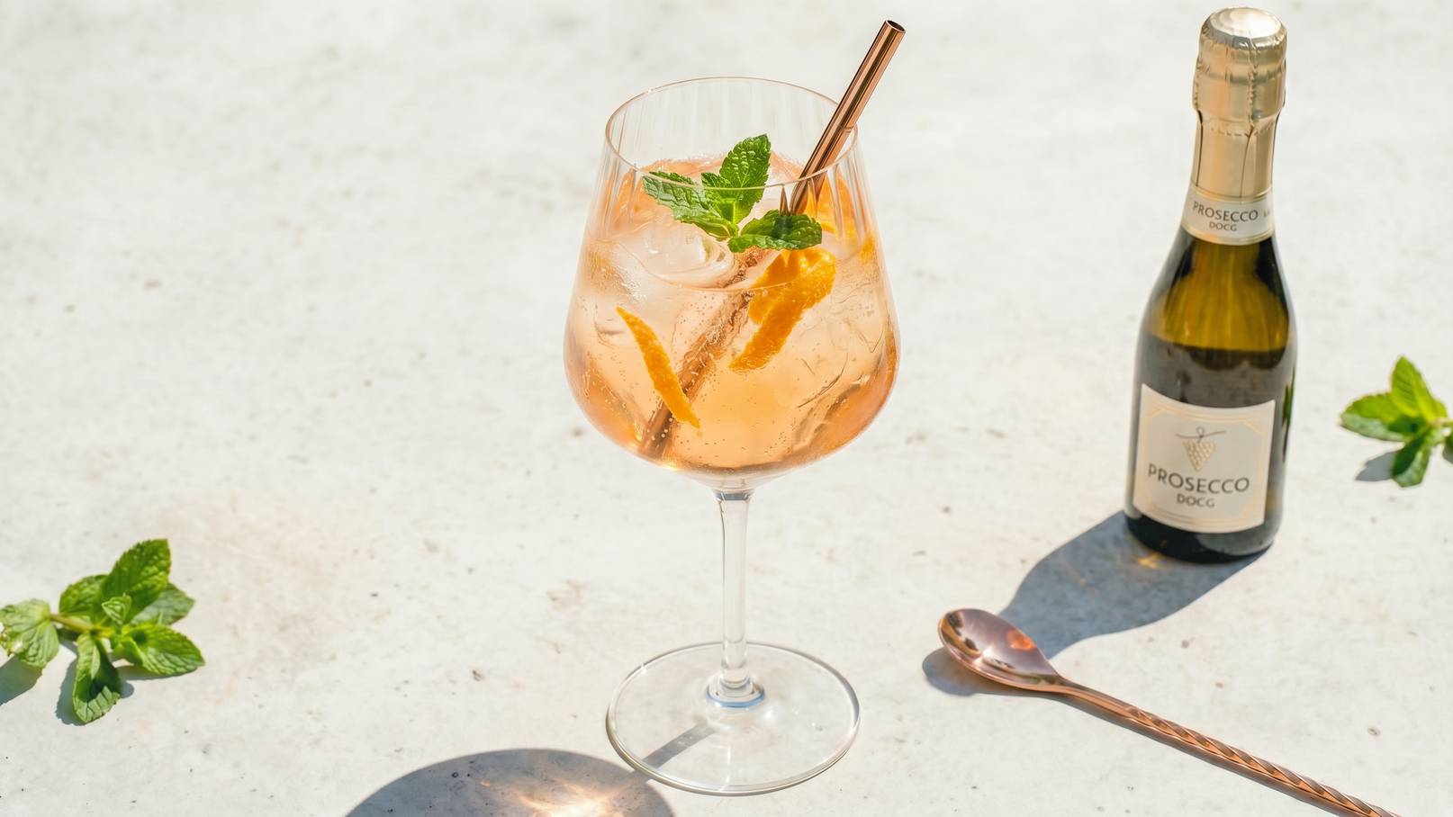 Ein erfrischender, rosafarbener Cocktail mit Eis, garniert mit Minzblättern und Orangenscheiben, steht in einem großen Weinglas. Neben dem Glas befindet sich eine kleine Flasche Prosecco und ein Cocktaillöffel auf einer hellen Oberfläche.