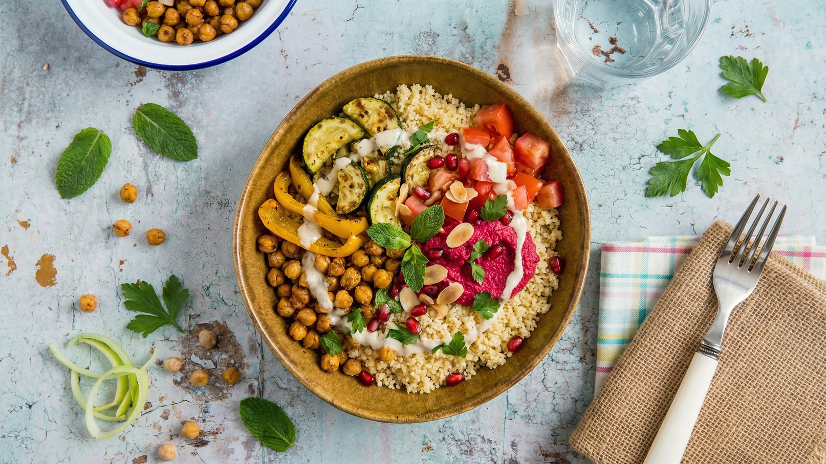 Eine Schüssel mit Couscous, garniert mit gegrilltem Gemüse, Kichererbsen, frischen Tomaten, Granatapfelkernen und einem Dip aus Roter Bete. Frische Kräuter wie Minze und Petersilie zieren das Gericht. Im Hintergrund befindet sich ein Glas Wasser und eine Gabel auf einem karierten Serviette.