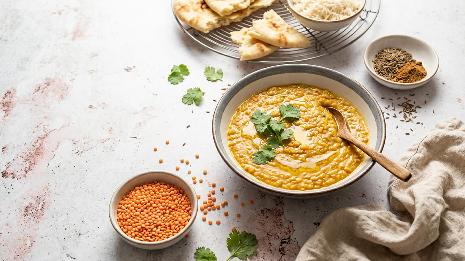 Eine Schüssel mit cremigem Linsencurry, garniert mit frischem Koriander und einem Holzlöffel. Daneben liegen eine Schale mit roten Linsen, einige Gewürze und ein Teller mit Brot. Im Hintergrund befindet sich eine weitere Schüssel mit Reis.