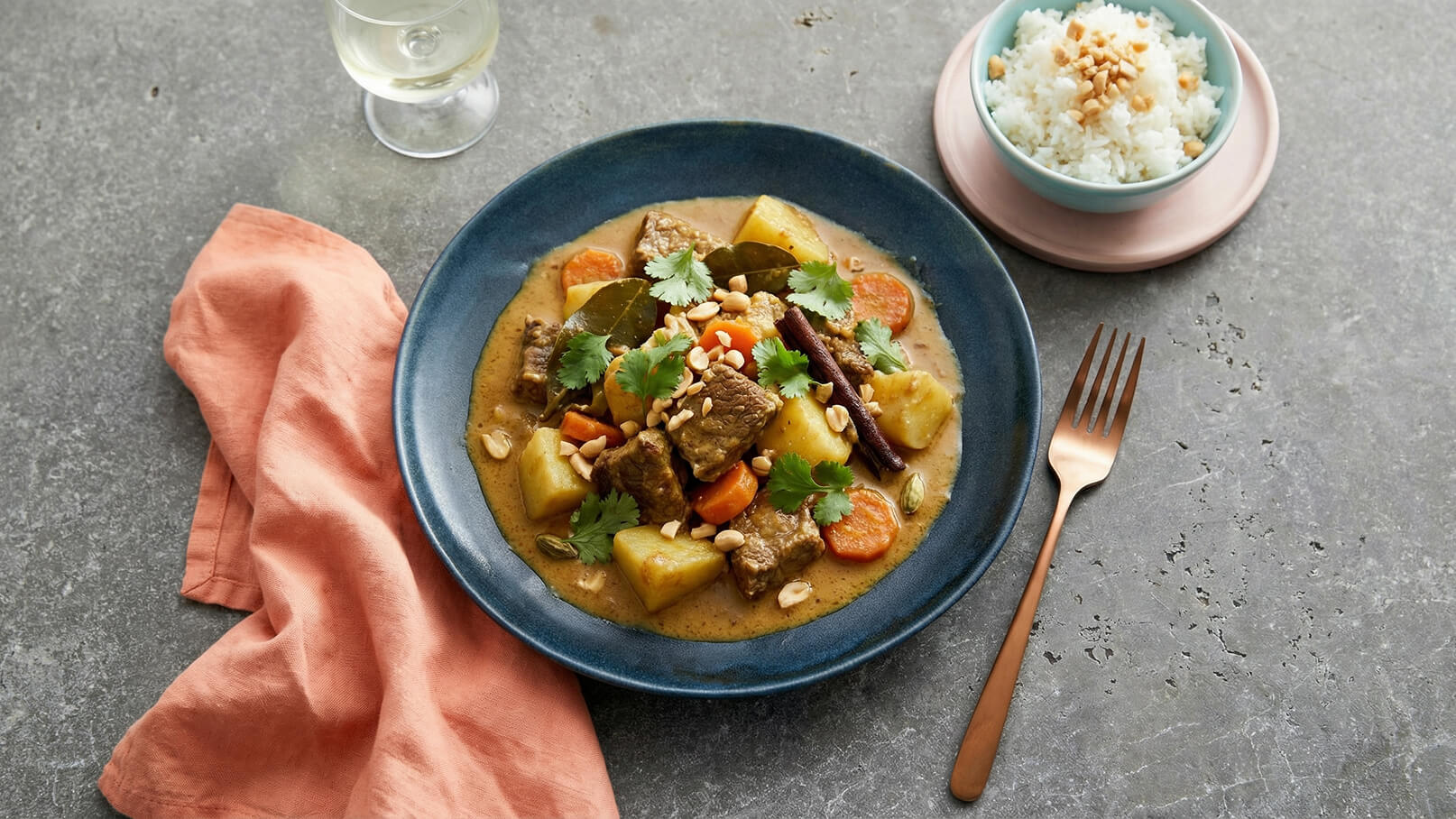 Ein blaues Tellergericht mit einer herzhaften Mischung aus Fleisch, Kartoffeln, Karotten und Gewürzen, garniert mit frischen Korianderblättern und Erdnüssen. Daneben ein kleiner Teller mit weißem Reis. Im Hintergrund steht ein Glas Weißwein. Ein oranges Tuch liegt daneben.