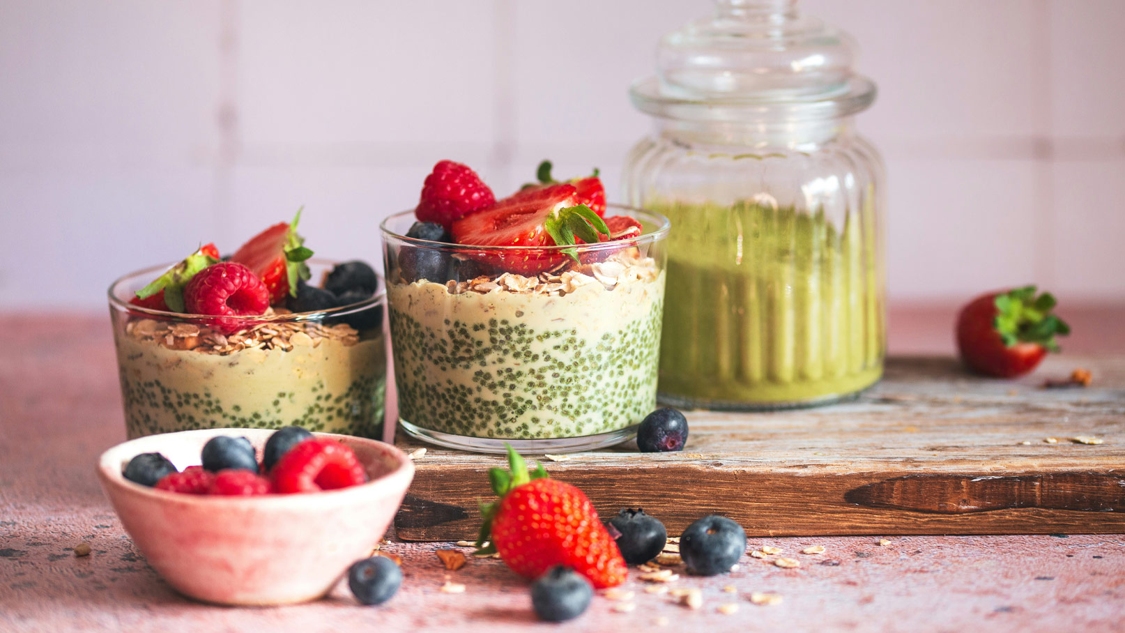Zwei Gläser mit Schichten aus Chia-Pudding und einer cremigen Füllung, dekoriert mit frischen Erdbeeren, Himbeeren und Blaubeeren. Neben den Gläsern steht eine Vorratsdose mit grünem Inhalt, während eine kleine Schüssel mit Beeren auf dem Tisch platziert ist.
